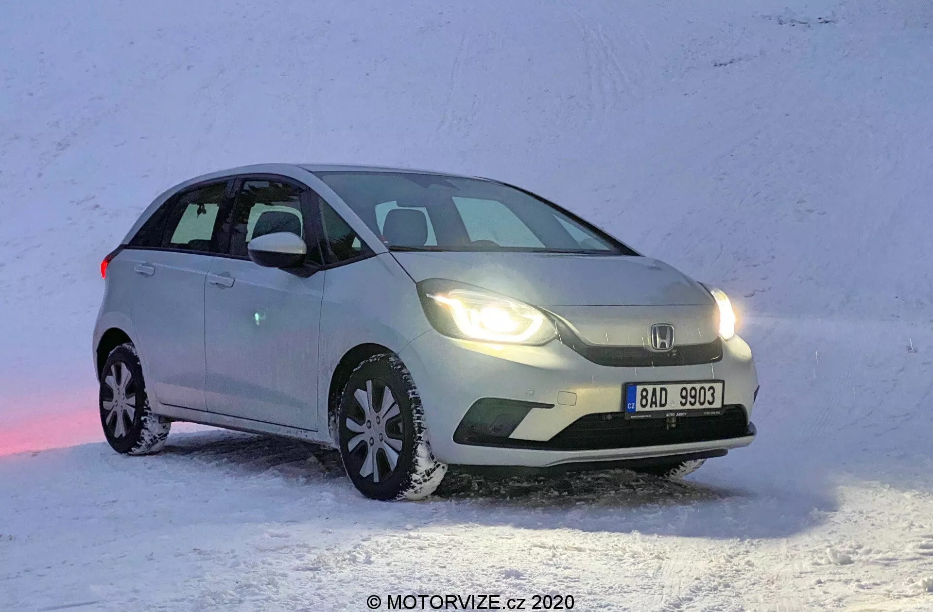 Uma vista frontal de três quartos de um Honda Jazz 2020 hatchback de cor clara, posicionado em uma superfície nevada. A imagem captura o carro do lado frontal esquerdo, mostrando o lado do motorista em um ângulo que destaca tanto o perfil lateral quanto a fachada frontal. O Honda Jazz está estacionado com a frente ligeiramente voltada para a direita, revelando o design da grade, dos faróis e parte do capô. Os faróis dianteiros estão acesos, refletindo na neve, com o veículo aparentemente parado. Pneus de neve com padrões de rodagem distintos são visíveis, destacando sua adequação para condução no inverno.