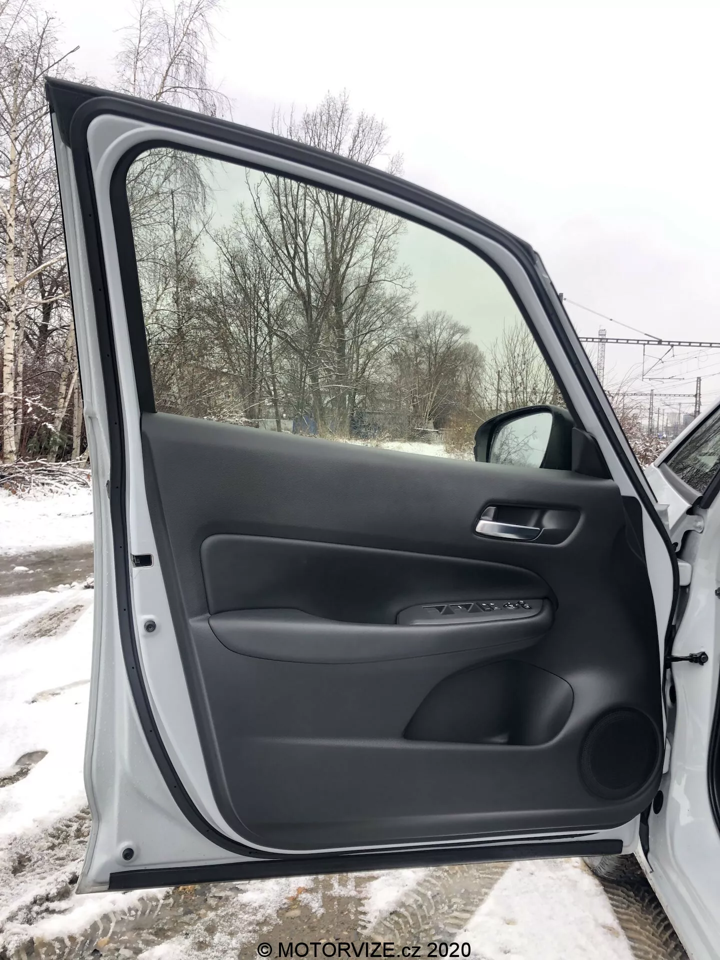 Vista interna da porta dianteira do passageiro do Honda Jazz 2020, totalmente aberta, apresentando materiais plásticos pretos texturizados e lisos. O painel da porta inclui um apoio de braço ergonômico, uma alça de material macio e um painel de controle para mecanismos de janela e travamento elétricos. Também há um detalhe prateado e um bolso de armazenamento inferior. Através da janela, vê-se uma paisagem nevada, indicando um cenário invernal.