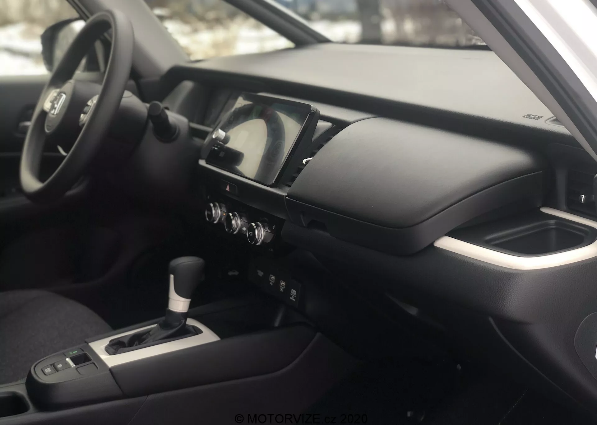 Vista interior do Honda Jazz 2020 do lado do passageiro, focando no painel e no cockpit do motorista. A fotografia, tirada em um ângulo ligeiramente elevado, destaca a console central, o volante multifuncional com controles montados, o painel de instrumentos digital-analógico, a console central com alavanca de câmbio, os botões de controle do clima e uma tela de infotainment/navegação. O apoio de braço sugere conforto para o motorista, e o interior possui materiais metálicos e de toque suave, refletindo uma filosofia de design moderna e de alta qualidade.