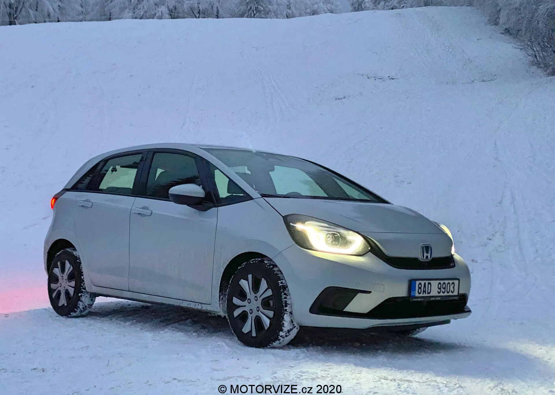 A imagem mostra um Honda Jazz 2020, um hatchback prateado ou cinza claro estacionado em uma superfície nevada, sugerindo uma cena de inverno com paisagem coberta de neve. Capturado de um ângulo frontal de três quartos, a vista destaca o lado do motorista, mostrando a grade frontal, faróis, capô, para-brisa, porta do motorista e porta traseira do passageiro. O carro apresenta faróis modernos, um para-choque dianteiro contornado, um emblema proeminente na grade, espelhos laterais com indicadores de seta integrados e rodas de liga leve multifusos equipadas com pneus de inverno. O veículo parado é mostrado sem desfoque de movimento sob possíveis condições de baixa luminosidade, seja no início da manhã ou no final da tarde.