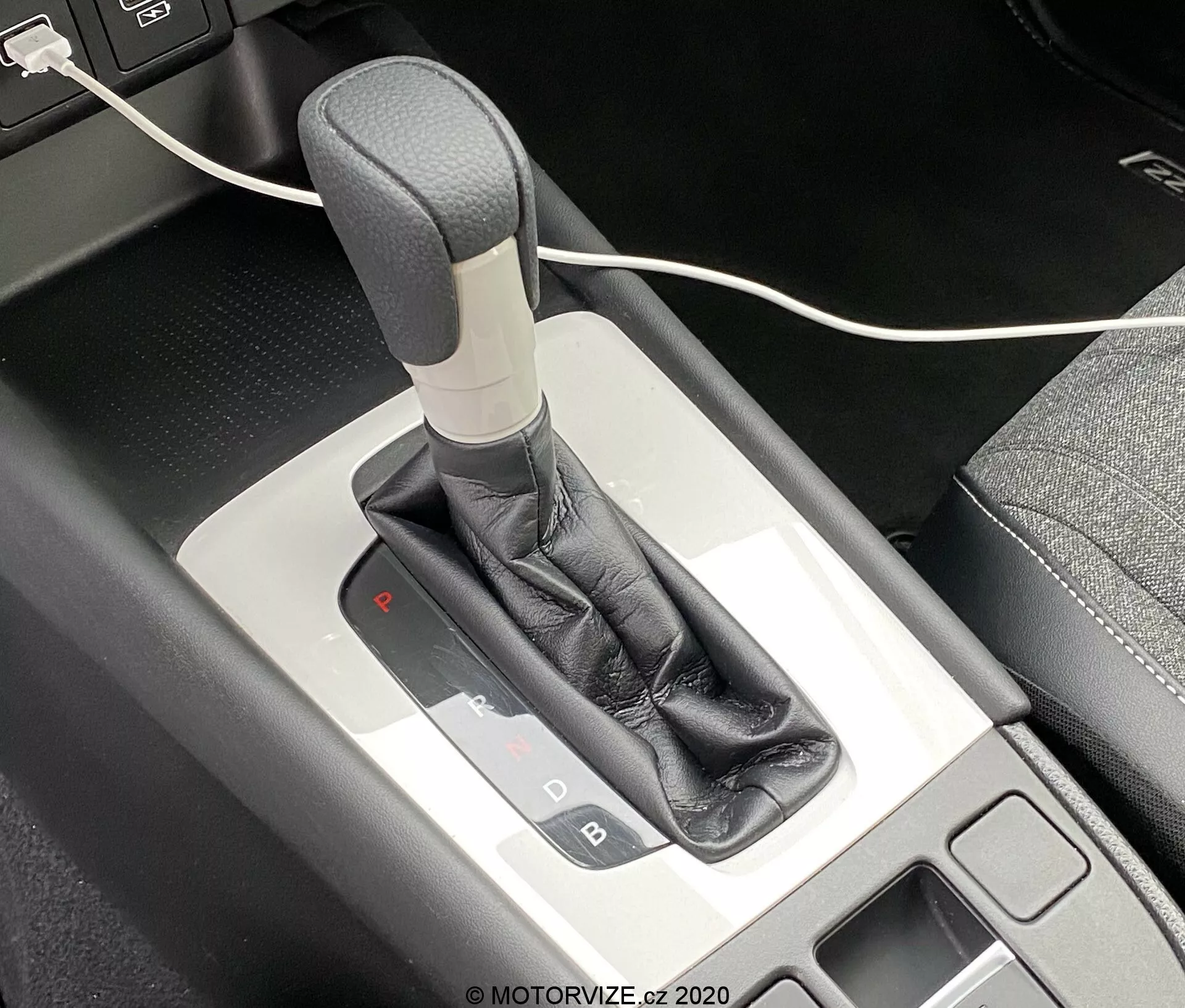Imagem de close-up da alavanca do câmbio automático do Honda Jazz 2020 a partir de um ângulo de cima para baixo, mostrando o mecanismo do seletor de marchas e console central ao redor. A alavanca de câmbio possui um manípulo cinza ou prateado, envolto em um revestimento de couro preto, complementado por um acabamento metálico. O painel de seleção de marchas iluminado indica que o veículo está no modo de estacionamento com uma luz vermelha sob o 'P'. Uma pequena área de armazenamento com um tapete preto é visível à esquerda, junto com um cabo USB parcialmente visível na parte superior da imagem.