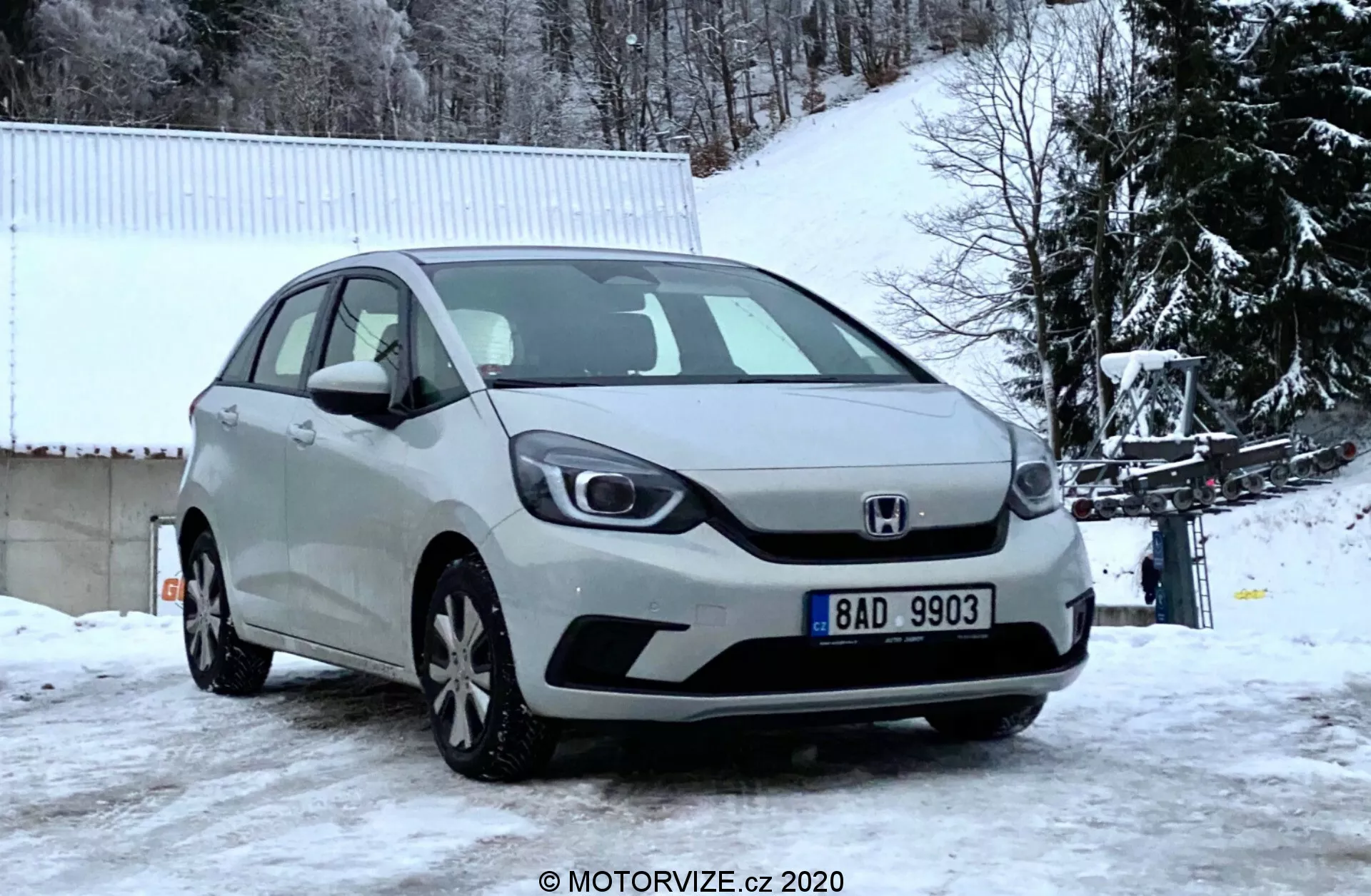 Vista de ângulo frontal de um Honda Jazz prateado de 2020 capturado em um dia de inverno, mostrando a grade frontal, faróis, capô e para-choque dianteiro contra um cenário coberto de neve com árvores nuas e um prédio branco.
