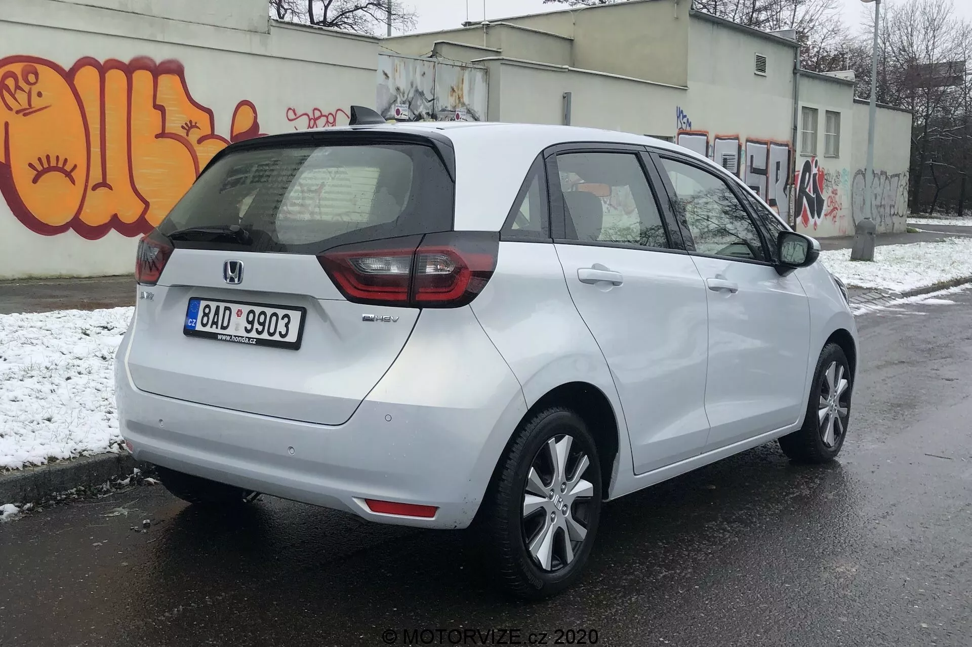Vista traseira de três quartos de um Honda Jazz 2020 branco estacionado em uma superfície asfaltada em um ambiente urbano potencialmente frio com neve no chão e grafite em uma parede. O carro possui uma combinação de luzes traseiras LED e tradicionais, placa de licença traseira central e um emblema indicando o nível de acabamento ou variante. Tem um teto aerodinâmico, rodas de liga leve de cinco raios, espelhos laterais na cor da carroceria com indicadores de mudança de direção integrados e maçanetas combinando com a cor da carroceria. O para-brisas traseiro está equipado com um desembaçador e limpador para melhor visibilidade.
