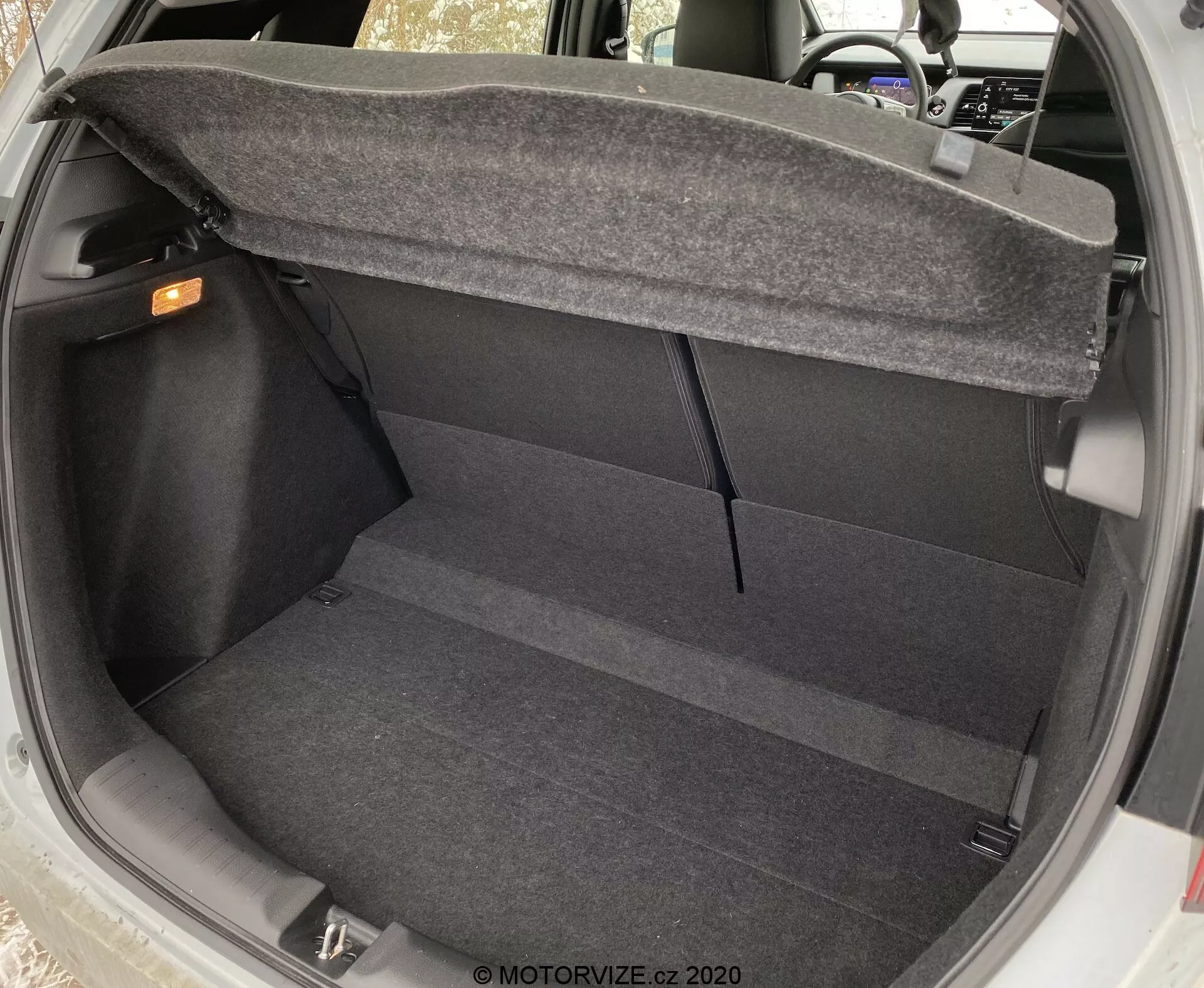 Vista interna do espaço do porta-malas do Honda Jazz 2020, a partir de um ângulo ligeiramente elevado, mostrando o piso limpo e acarpetado, as paredes laterais e a parte de trás dos bancos traseiros. O porta-malas está vazio, com a alça de liberação interna visível no canto superior direito e o mecanismo de dobradiça da tampa do porta-malas parcialmente visível nos lados superiores, destacando a praticidade e os recursos de design básicos do porta-malas.