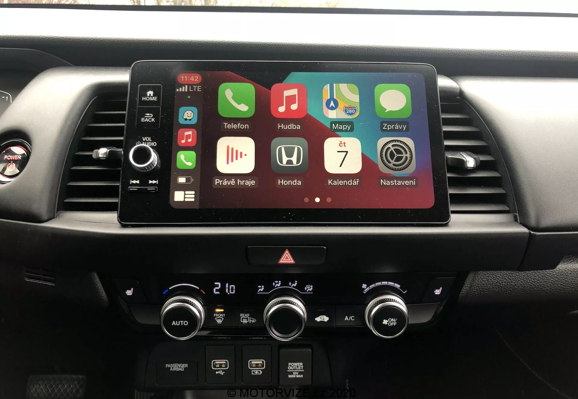 Console central do interior do Honda Jazz 2020, mostrando sistema de infotainment e controle do clima a partir da perspectiva do motorista. Tela touchscreen ativa com aplicativos como Telefone, Música, Mapas indicando Bluetooth, GPS, reprodução de mídia e Apple CarPlay. Abaixo da tela, botões físicos, luzes de alerta e controles de clima de zona dupla com leituras em Celsius.
