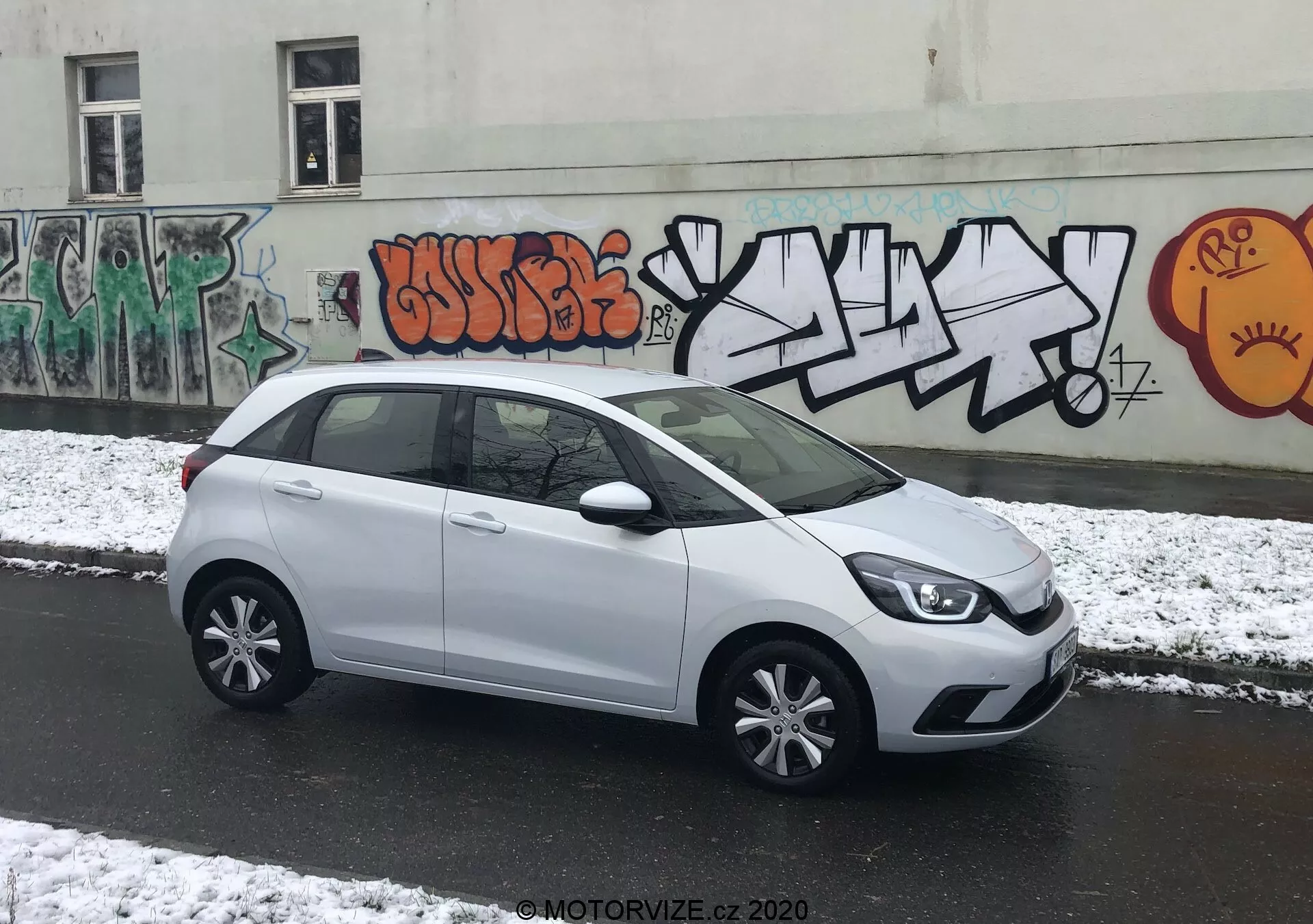 A imagem apresenta um hatchback Honda Jazz 2020 branco estacionado perpendicularmente à câmera em uma rua urbana, permitindo uma visão completa de seu perfil lateral. A frente elegante do carro com seus clusters de faróis modernos é parcialmente visível, levando a uma linha de caráter suave na lateral. As maçanetas das portas e espelhos laterais combinam com a cor da carroceria, mantendo uma estética coesa. A ampla área envidraçada oferece amplo espaço para a cabeça, e uma pequena janela lateral traseira adiciona ao design. O spoiler traseiro sugere eficiência aerodinâmica. A iluminação nublada com neve fresca e o cenário urbano com graffiti acrescentam um ambiente dinâmico.