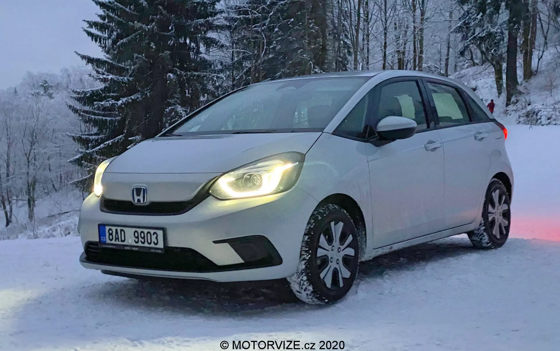 Fotografia profissional de um Honda Jazz branco de 2020 estacionado em terreno nevado durante o inverno. A imagem é tirada em um ângulo que foca o lado frontal esquerdo do carro, mostrando a grade frontal, faróis de LED, capô, roda dianteira esquerda de liga leve com vários raios equipada com pneus de neve, para-brisa e porta do motorista. Árvores cobertas de neve são visíveis ao fundo, e a placa é legível. A luz fraca sugere início da manhã ou fim da tarde, com as luzes diurnas do carro refletindo na neve, realçando a ambientação da cena. O veículo possui iluminação avançada e setas integradas nos retrovisores laterais, com o emblema distintivo da Honda exibido de maneira proeminente na grade.