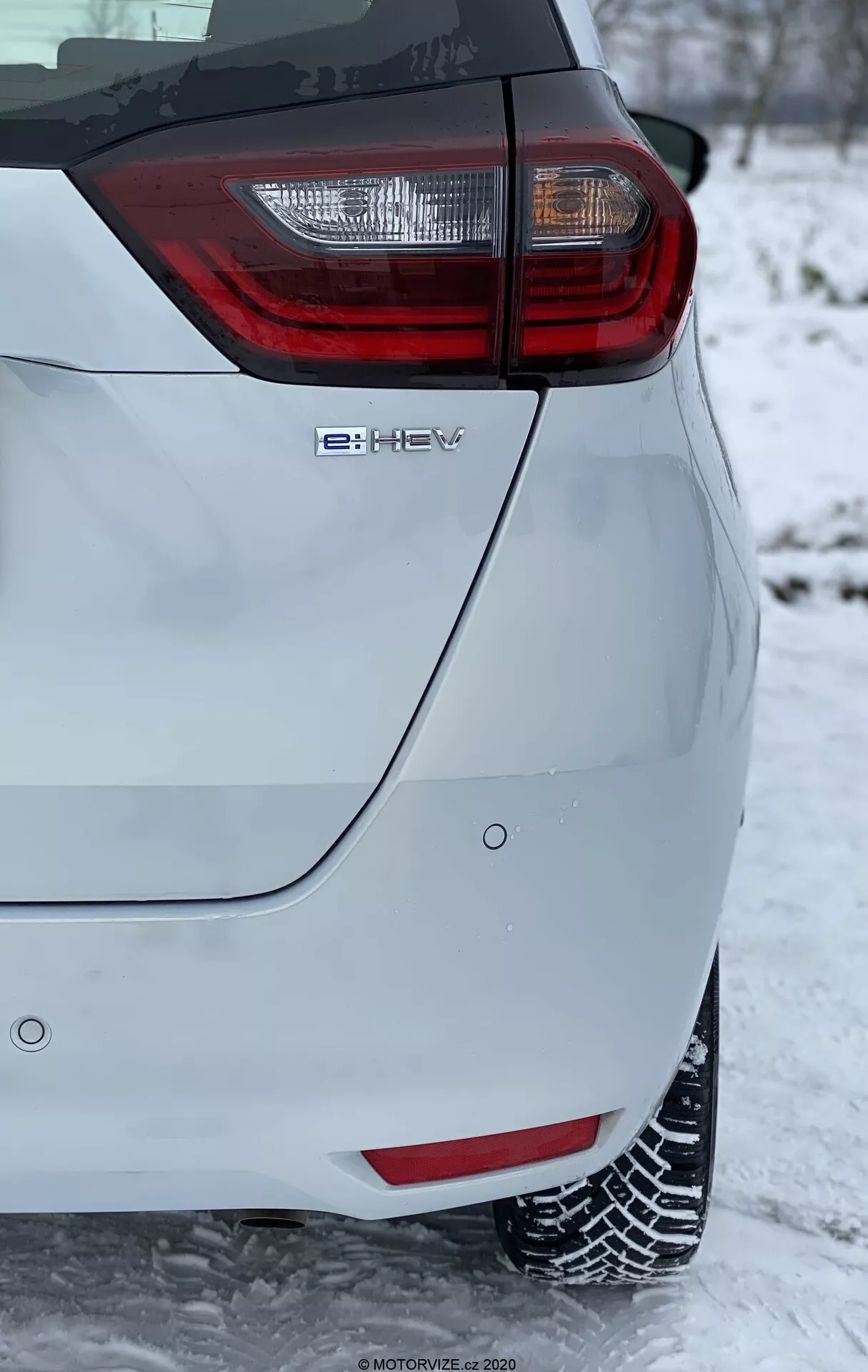 Close-up do canto traseiro de um carro hatchback moderno, com um proeminente conjunto de luzes traseiras com elementos vermelhos e claros. A imagem, tirada em orientação vertical, mostra o emblema de tecnologia híbrida 'e-HEV' e está ambientada em um ambiente de inverno com neve no chão e cobrindo os pneus, indicando condições de condução de inverno. A pintura de cor clara do veículo contrasta com o fundo nevado, destacando suas características de design moderno e ecológico.