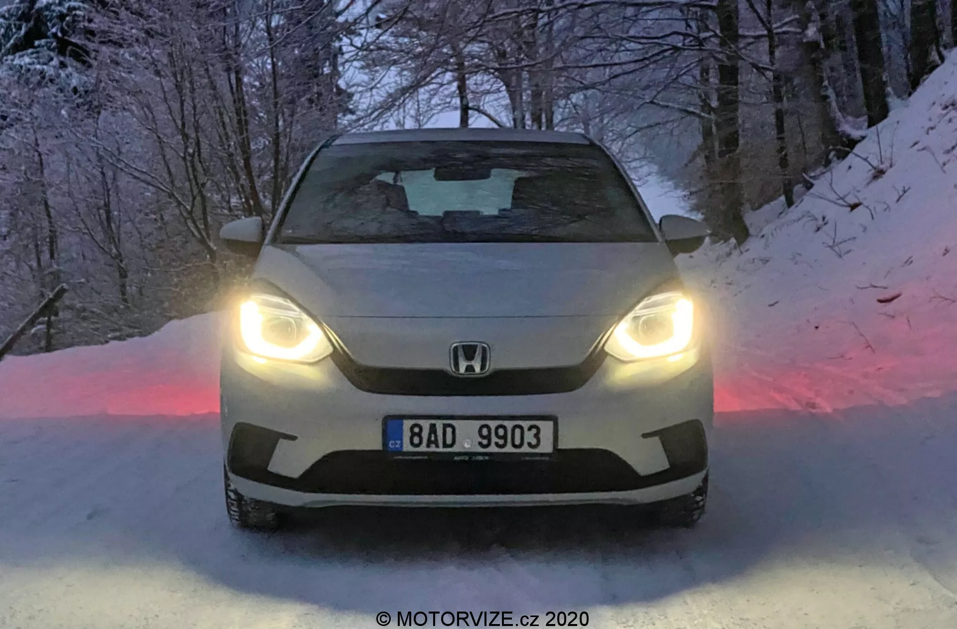 Imagem frontal do Honda Jazz 2020 iluminado com faróis LED, mostrando o emblema da marca e a placa na grade, com capô esculpido e para-brisa escurecido. O chão coberto de neve e o crepúsculo realçam o design avançado e a capacidade de condução no inverno do carro.