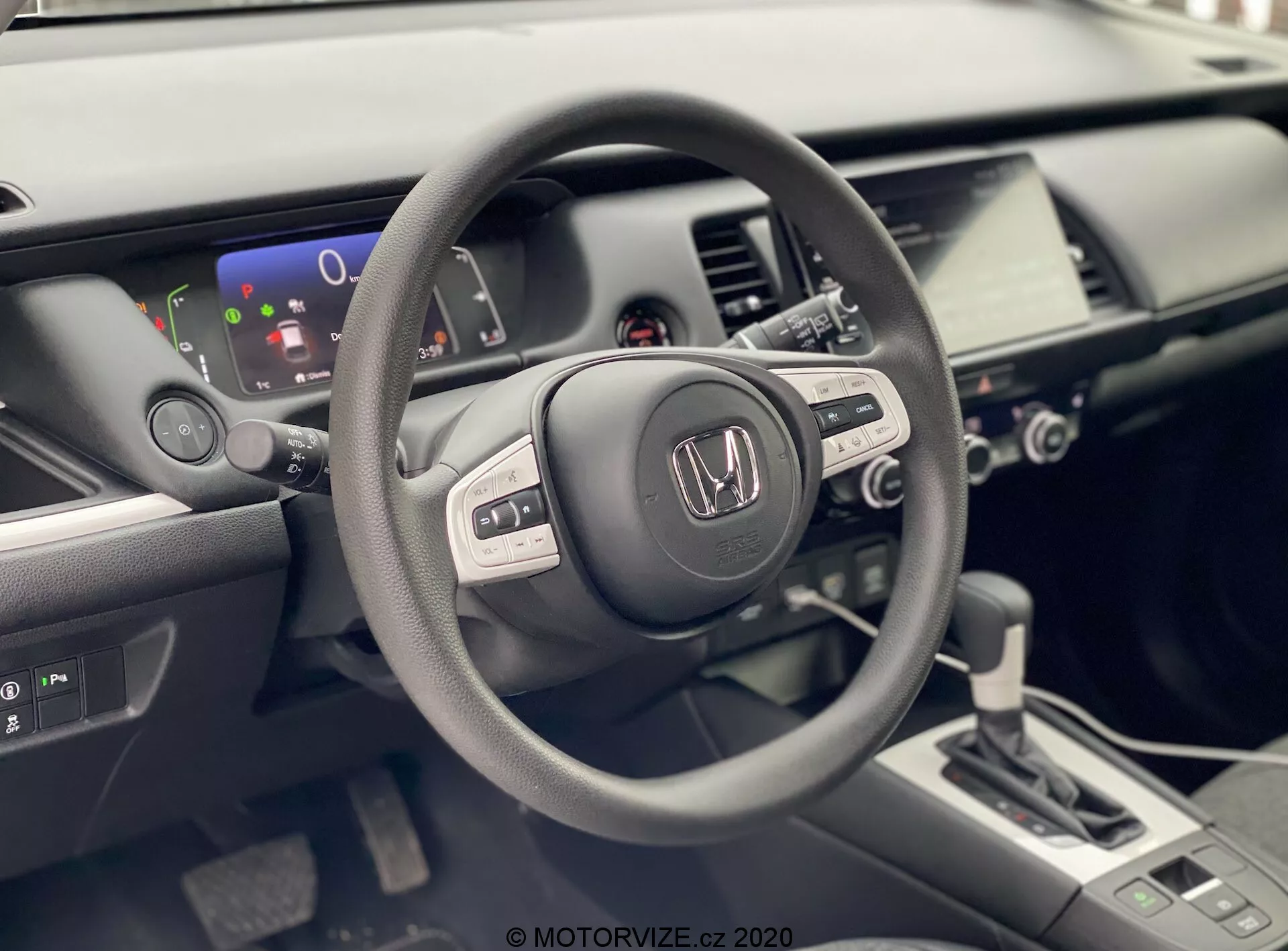 A foto mostra o interior de um Honda Jazz 2020, visto da perspectiva do motorista. O foco está no volante com a marca Honda, que possui vários botões de controle sugerindo funcionalidades integradas de áudio, display de informações e possivelmente controle de cruzeiro. Atrás do volante, é visível um display digital parcial que mostra um indicador de nível de bateria, sugerindo que o veículo pode ser híbrido ou totalmente elétrico. O interior bem iluminado e moderno também inclui um console central com um sistema de infotainment touchscreen, controles de climatização e uma alavanca de câmbio de design contemporâneo indicando uma transmissão automática. A imagem destaca os acabamentos em mate, acetinado e brilhante usados nos materiais interiores do carro, enfatizando o design ergonômico e rico em tecnologia do veículo.