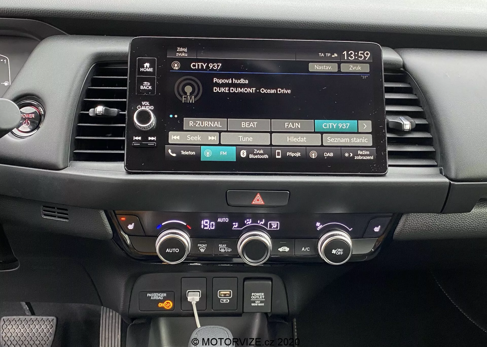 Close-up da área central do painel de controle do Honda Jazz 2020. A imagem apresenta proeminentemente o display de toque do sistema de infotainment, rodeado por botões e manípulos físicos para ajustes de áudio, mídia e configurações do veículo. Abaixo, há duas ventilas de ar e um sistema de controle climático abrangente com botões e manípulos para ajuste de temperatura, velocidade do ventilador, funções de desembaçamento e recirculação. O painel é composto por materiais modernos, incluindo plásticos foscos e brilhantes, projetado com acessibilidade funcional e facilidade de uso em mente.