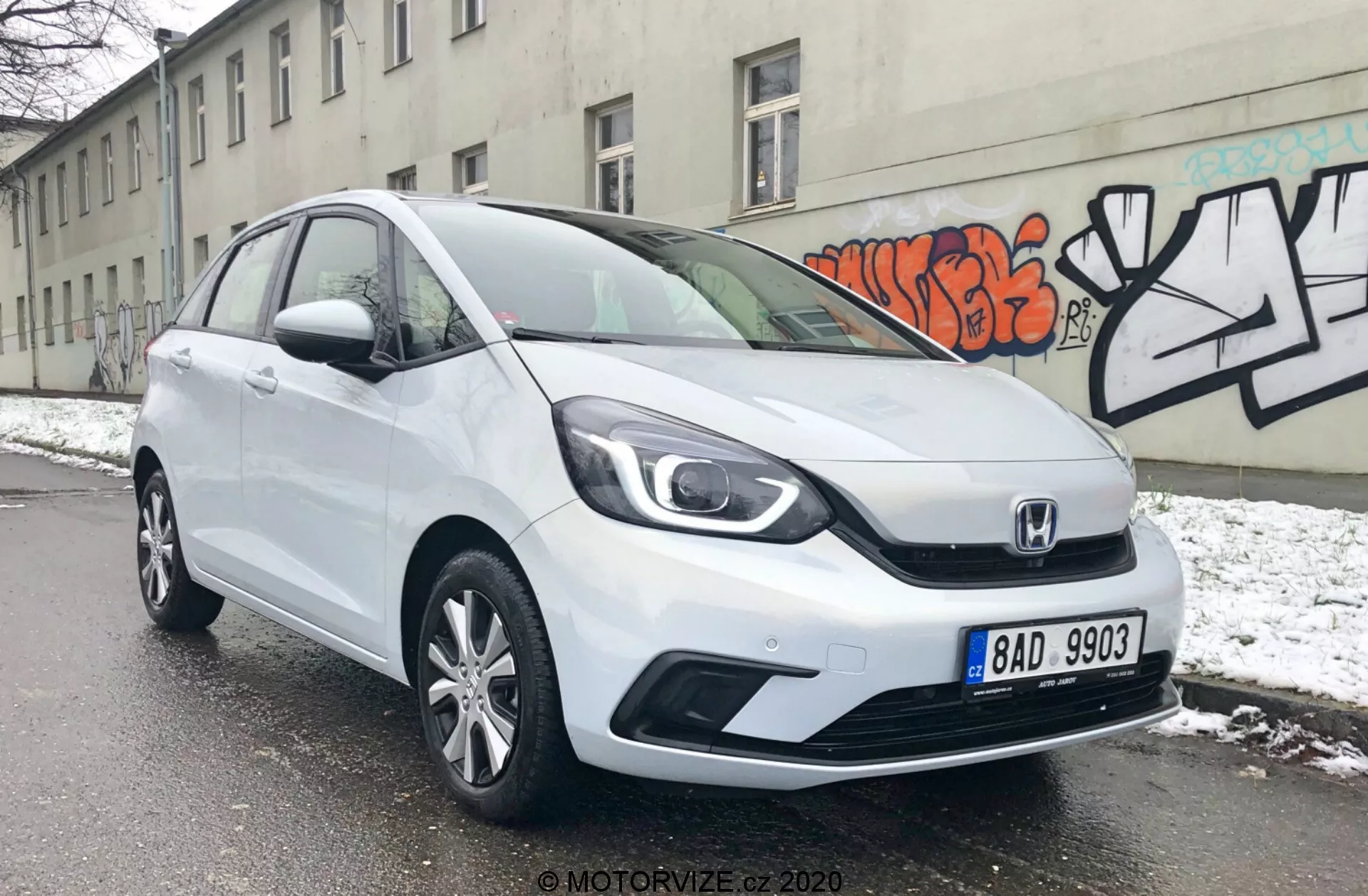 A imagem mostra um Honda Jazz 2020 hatchback branco em um ângulo de três quartos frontais, revelando seus elegantes faróis, a grade distintiva da Honda com um design de duas barras e as linhas de contorno aerodinâmicas no capô. O para-choque dianteiro apresenta entradas de ar visíveis e possivelmente luzes de neblina, enquanto o perfil lateral exibe rodas de liga leve multirraios e painéis de porta ligeiramente contornados. O carro está estacionado em uma rua nevada, indicando um ambiente de inverno, com uma parede cinza decorada com grafites ao fundo, destacando um cenário urbano.