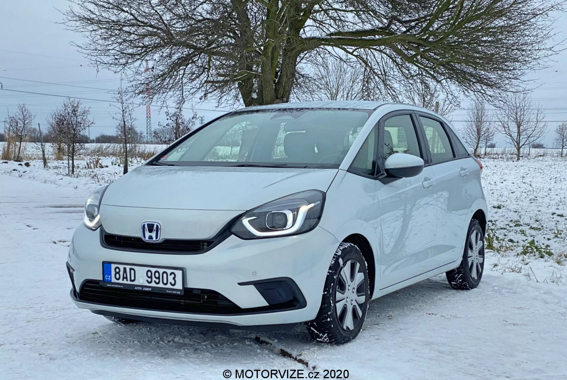 Vista em ângulo frontal de três quartos de um Honda Jazz 2020, mostrando a parte frontal e parte do lado do passageiro deste moderno hatchback compacto. A imagem, aparentemente tirada na altura dos olhos, destaca a grade minimalista com o emblema central da Honda, faróis elegantes com luzes diurnas de LED, rodas de liga leve multi-raio e espelhos laterais com indicadores de direção integrados. O carro está estacionado em uma área pavimentada parcialmente coberta de neve, sugerindo um cenário de inverno com árvores sem folhas e um céu cinza ao fundo. O veículo parece limpo e bem conservado.