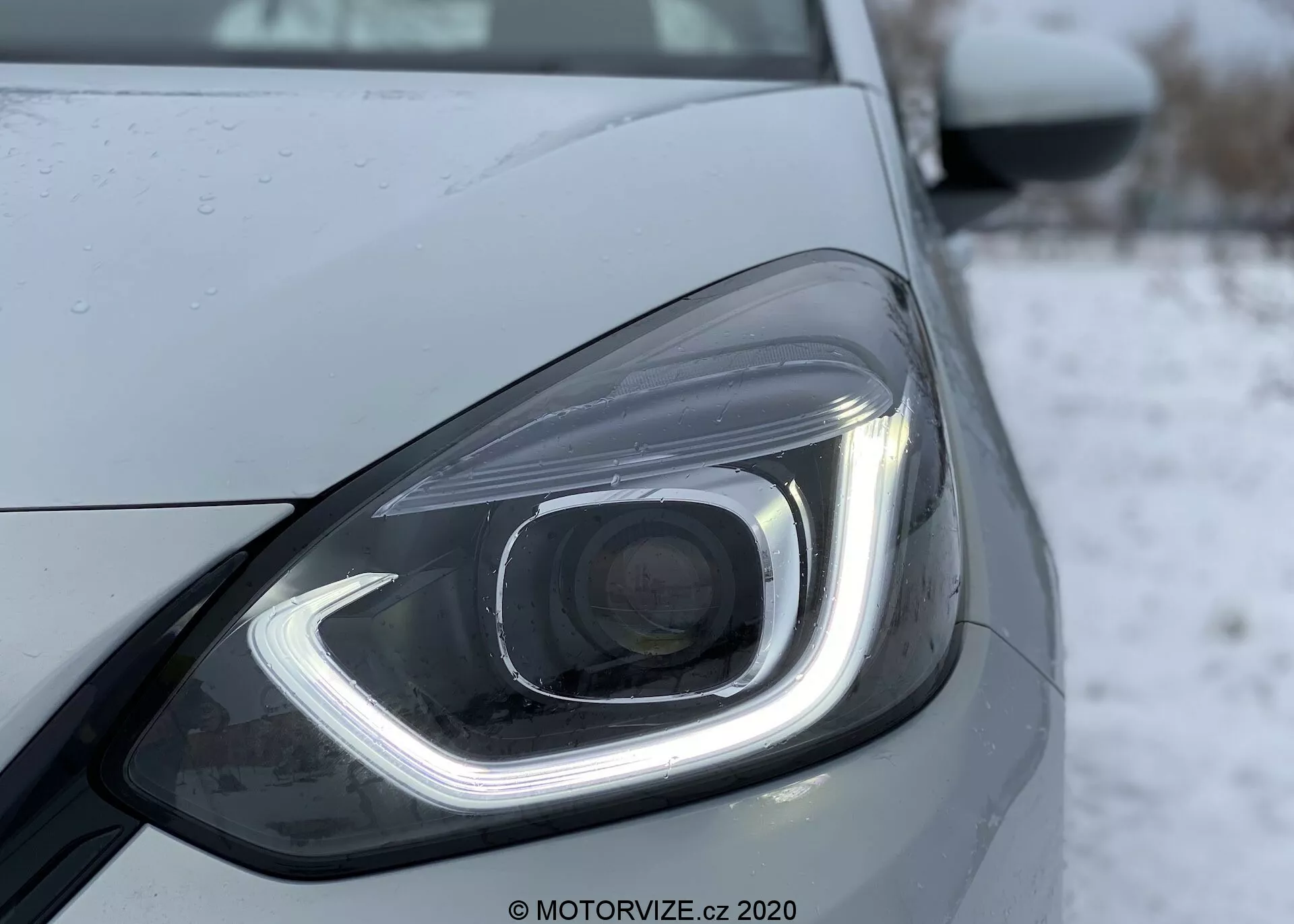 Fotografia em close-up do farol dianteiro esquerdo de um Honda Jazz 2020, destacando seu design moderno com elementos de iluminação LED ou HID. O farol apresenta uma luz diurna distintiva em forma de gancho, uma lente projetora e gotas de água que sugerem precipitação recente ou lavagem. O design elegante é enfatizado por linhas e contornos suaves, com um fundo frio ou invernal, possivelmente nevado.