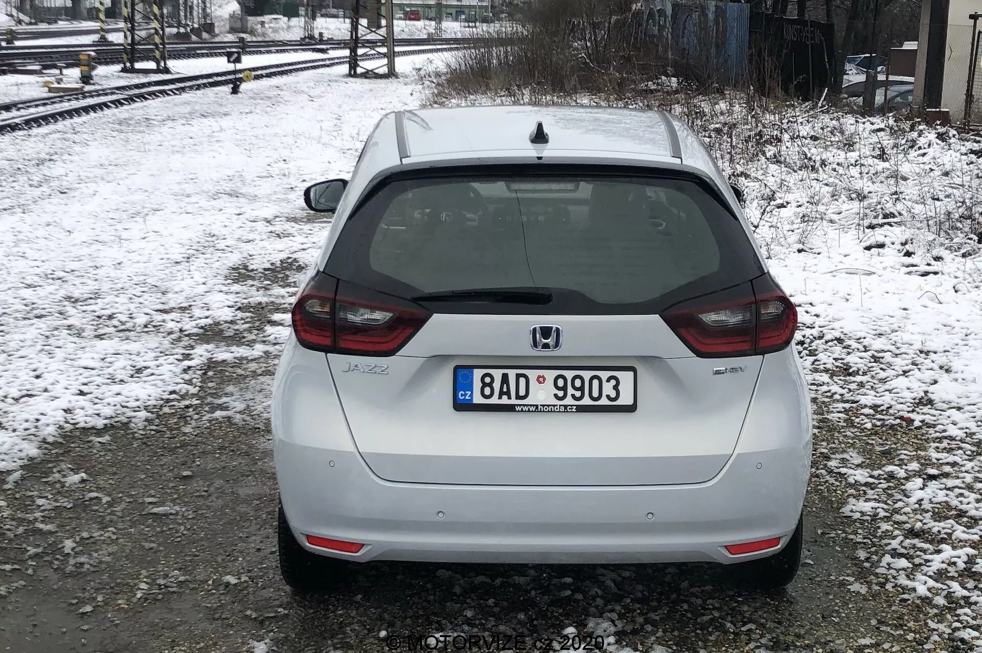 A imagem mostra a vista traseira de um Honda Jazz 2020 branco, estacionado em um chão nevado perto de trilhos de trem sob condições de luz difusa ou nublada. O carro ocupa a maior parte do quadro, com uma visão traseira centralizada que mostra sua placa de estilo europeu, limpador de para-brisa traseiro, luz de freio montada no alto e o emblema da Honda. O emblema do modelo 'JAZZ' é visível na parte inferior esquerda do porta-malas, e há uma câmera de ré acima da placa. As lanternas traseiras envolvem parcialmente as laterais, com refletores na parte inferior do para-choque, que também tem uma seção preta integrada. Os sinais de sujeira sugerem que o carro foi conduzido em estradas molhadas ou lamacentas. A neve e as árvores nuas indicam que é inverno ou início da primavera.