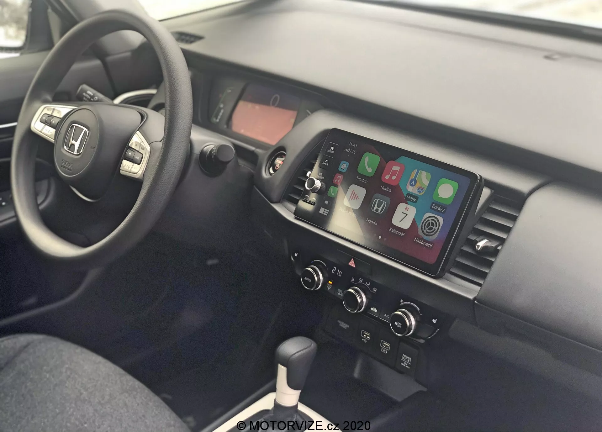 Vista interior de um Honda Jazz 2020, mostrando a área do motorista e o console central a partir do lado do passageiro. O volante está à esquerda e possui controles montados para áudio, conectividade telefônica e controle de cruzeiro, indicando um design para dirigir pelo lado direito da estrada. O console central conta com uma tela sensível ao toque que mostra a integração com smartphone, como Apple CarPlay ou Android Auto, controles manuais de clima, alavanca de câmbio para transmissão manual, portas USB e uma tomada de 12V, enfatizando a conectividade moderna e o design funcional.