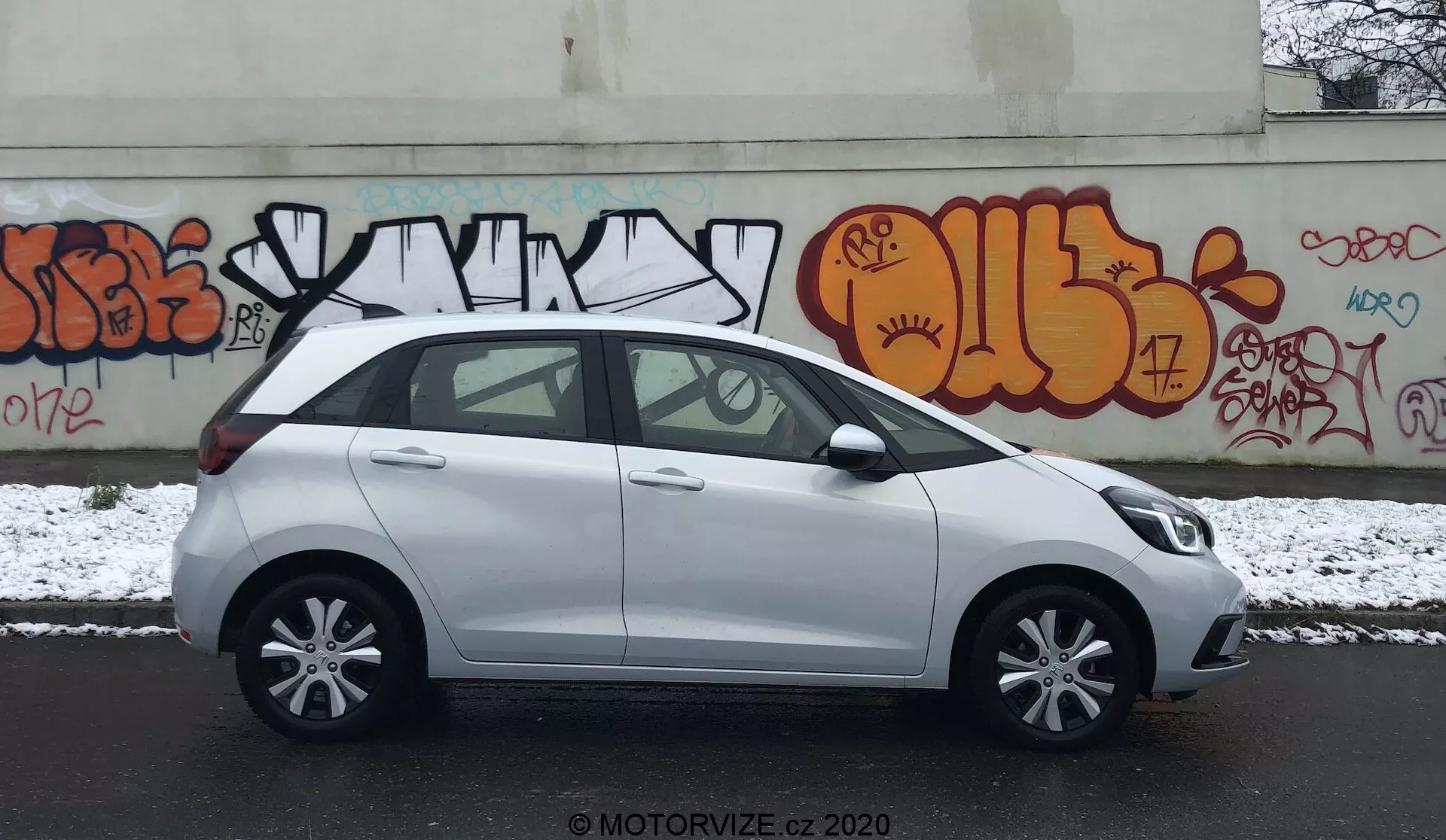 Perfil lateral de um Honda Jazz compacto branco de 2020 estacionado numa rua urbana com grafites na parede de fundo. A cena é num dia nublado com uma leve camada de neve no chão, sugerindo condições invernais. A imagem capta o lado do motorista, exibindo maçanetas de portas da cor do carro, espelhos laterais, jantes de liga leve de cinco raios e janelas padrão com um pilar B preto. O veículo aparenta estar limpo com algumas manchas úmidas da neve. A marca d'água 'MOTORVIZE.CZ 2020' é visível, indicando a fonte da foto.