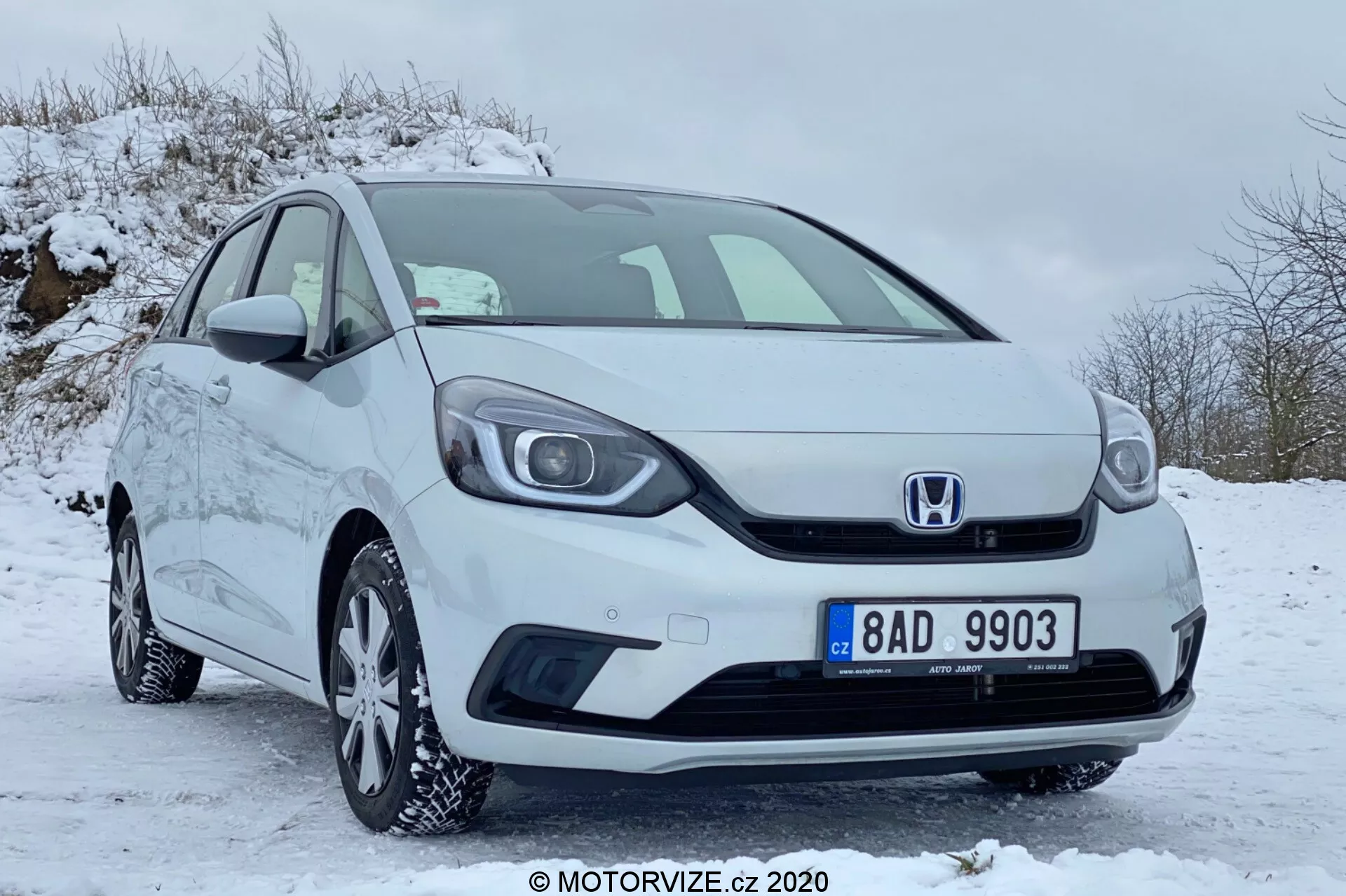 Vista frontal de três quartos de um carro compacto branco Honda Jazz 2020 estacionado em uma área coberta de neve, mostrando a grade com o emblema Honda, o para-choque dianteiro, os faróis, as jantes de liga leve com pneus de inverno, os espelhos laterais e as maçanetas das portas, tudo em condição bem mantida e limpa.