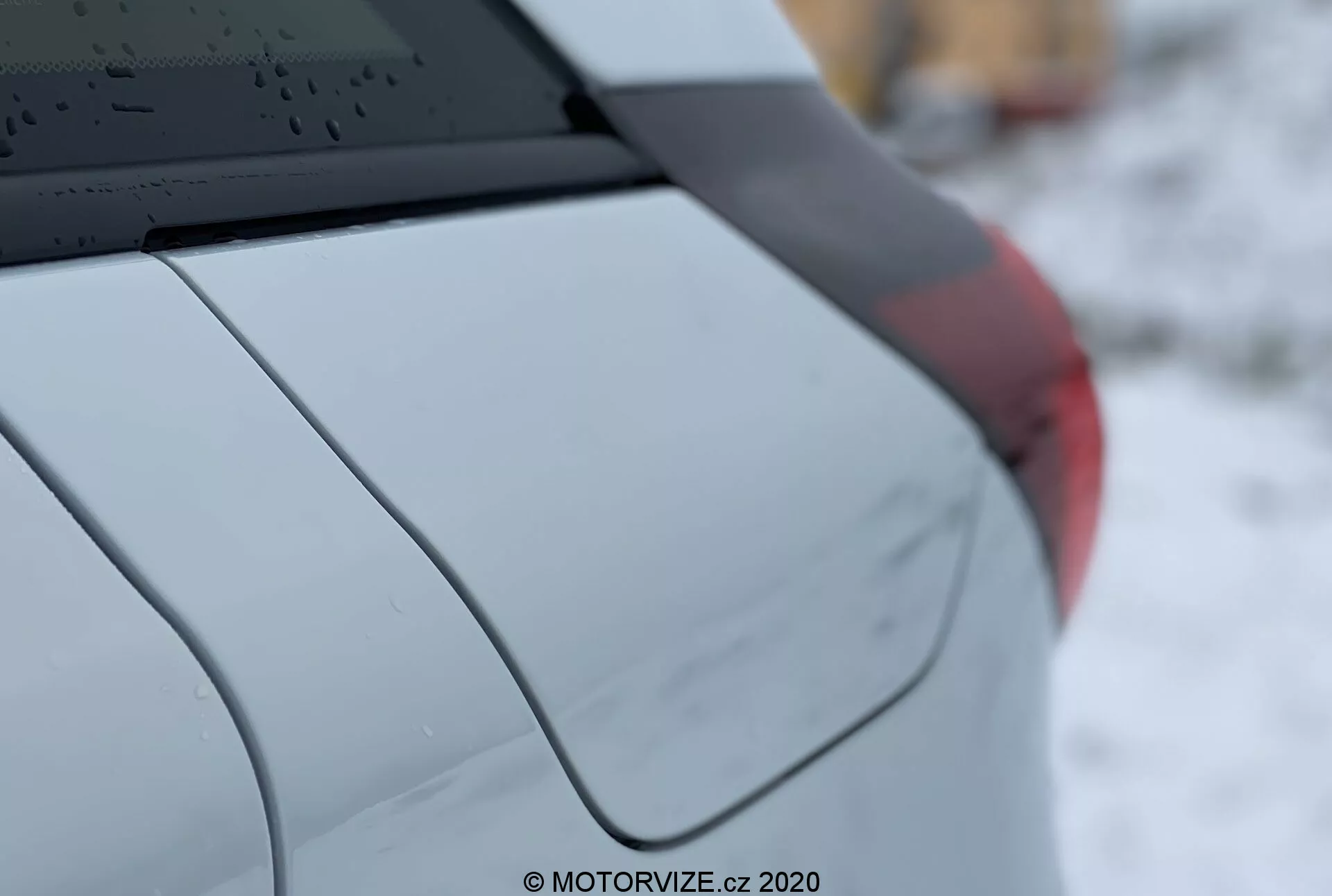 Detalhe em close-up do Honda Jazz 2020, mostrando o quarto traseiro direito do carro em uma vista lateral de três quartos. As características incluem um vidro traseiro com linhas descongeladoras, uma antena em forma de barbatana de tubarão montada no teto, o pilar C acentuando o design do carro, um para-lama traseiro suavemente curvado, um fragmento de uma unidade de luz traseira e um acabamento de pintura metálica ou perolada. A pequena profundidade de campo destaca os detalhes do veículo enquanto o fundo está desfocado. A marca d'água diz 'MOTORVIZE.cz, 2020.'