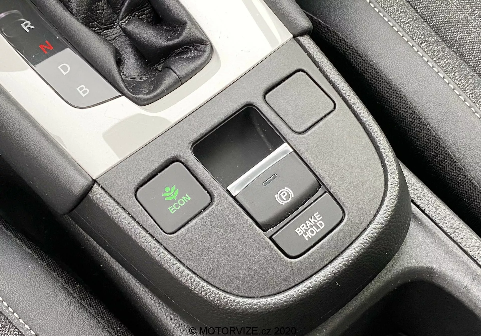 Close-up do console central interior de um Honda Jazz 2020 a partir de uma ligeira perspectiva superior do motorista. O seletor de marchas com 'N' para Neutro é parcialmente visível no canto superior direito, indicando transmissão automática. Destacam-se o botão ECON à esquerda para o modo econômico, um pequeno compartimento retangular de armazenamento ao centro e dois botões empilhados à direita: um com um ícone de energia e o outro rotulado 'BRAKE HOLD', que evita o deslocamento em paradas. O console possui acabamento premium com textura semelhante ao couro e detalhe de costura.