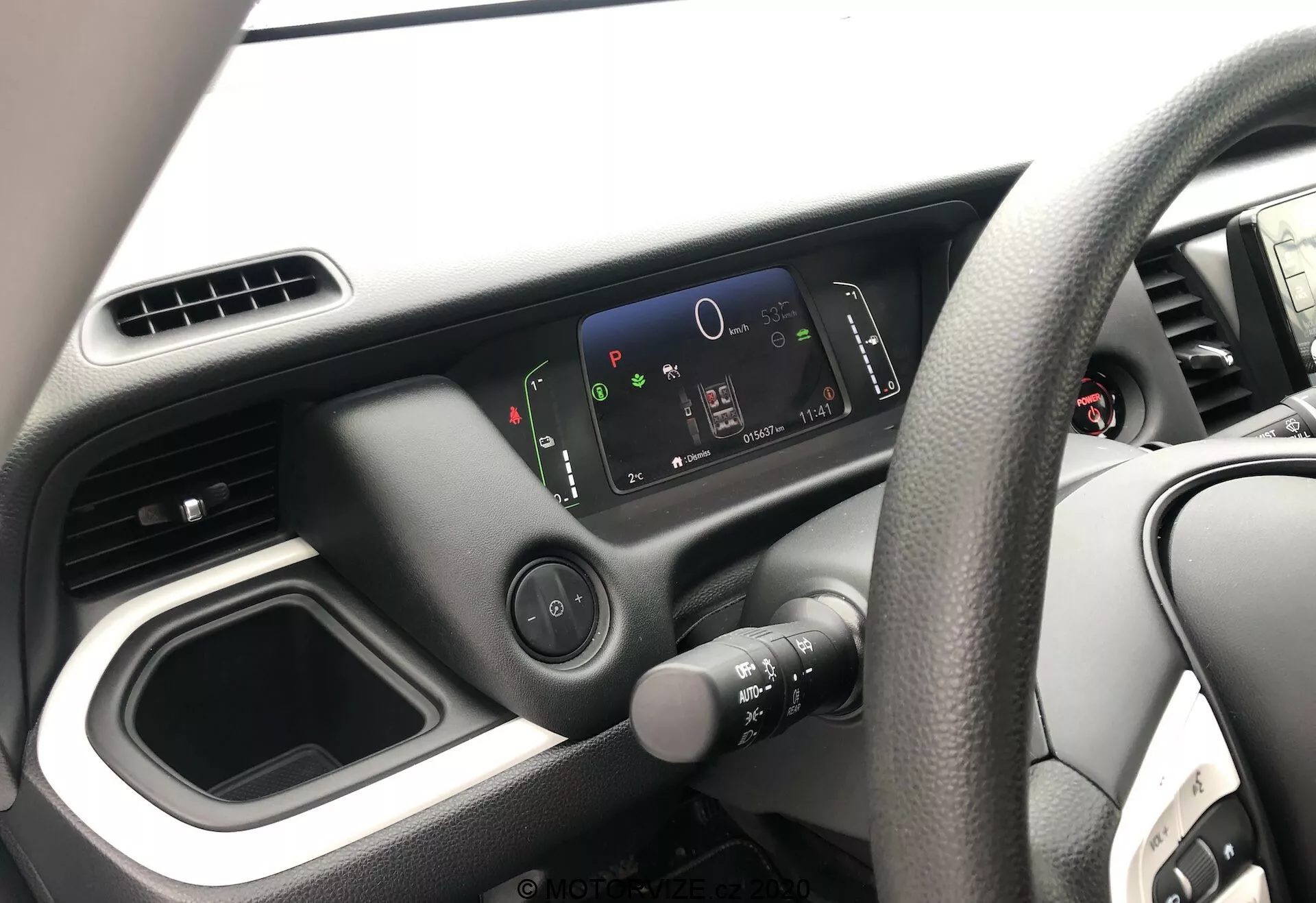 Vista interior do Honda Jazz 2020, focando no lado do motorista a partir da perspectiva do passageiro. A imagem mostra o volante à direita com design de quatro raios, o painel minimalista com um cluster de instrumentos digital de alta resolução e o layout simples do console central com um compartimento de armazenamento vazio e aberturas de ar. Apenas uma parte da porta do motorista está visível.