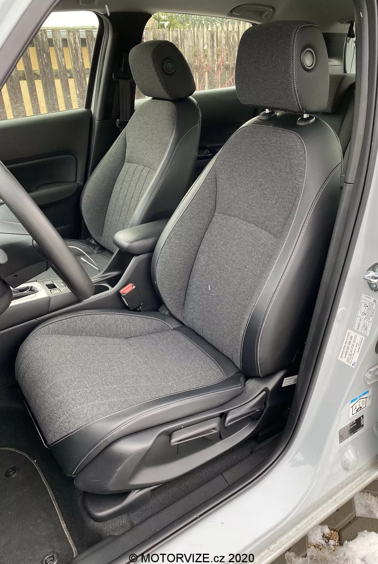 Imagem mostra o interior de um Honda Jazz 2020, focando na área do assento do motorista, visto de um ângulo ligeiramente acima da linha dos olhos do lado de fora do veículo. O assento ajustável do motorista é estofado em tecido escuro. São visíveis alavancas de ajuste para deslizar e reclinar o assento, o painel interno da porta com controles de vidros elétricos, o volante parcialmente visível com controles montados no volante, uma parte do painel, o tapete do piso e a área dos pedais indicando possível transmissão manual, parte do console central e o cinto de segurança do motorista pendurado ao lado.