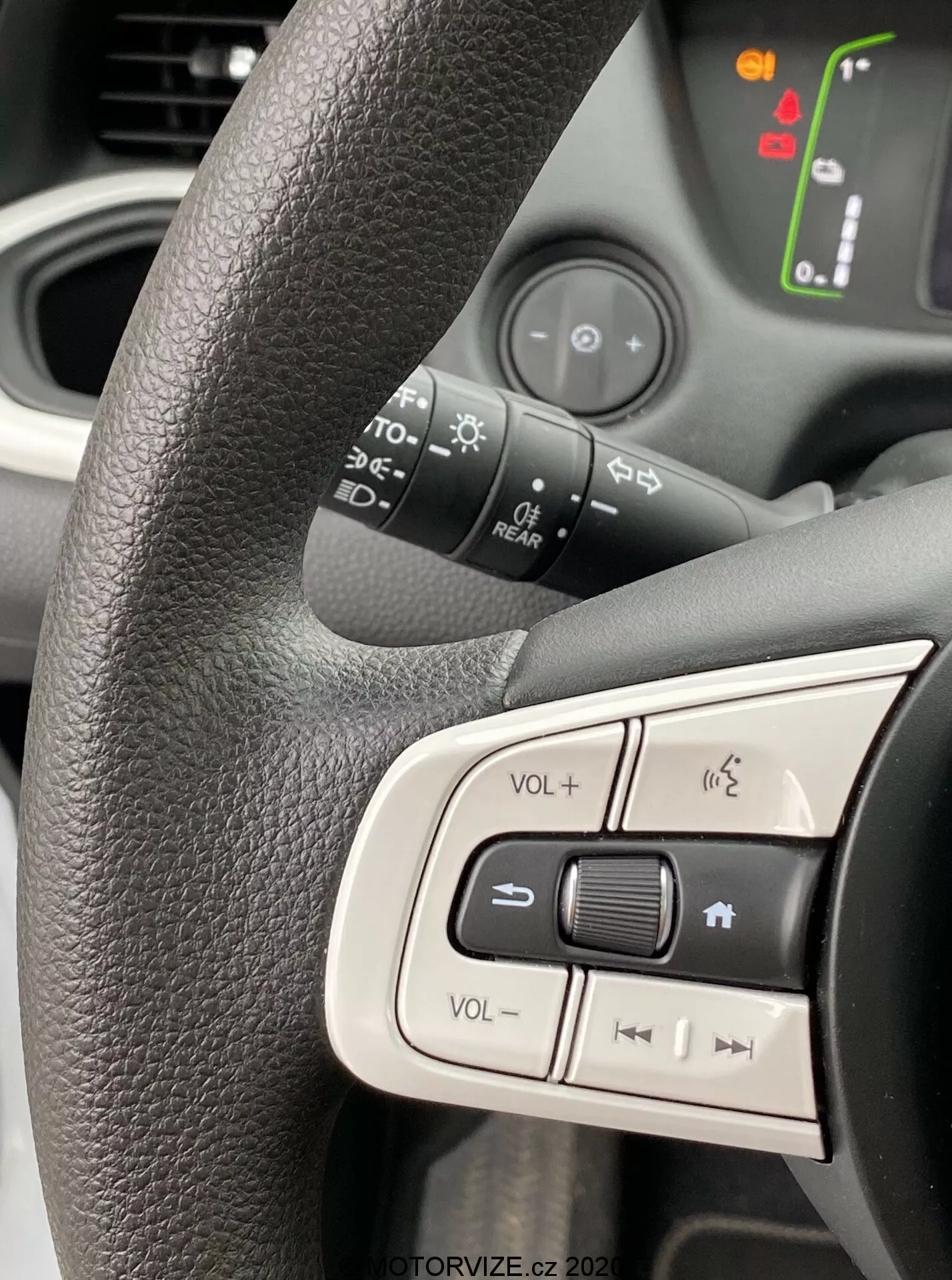 Visão de perto do lado esquerdo do volante de um Honda Jazz 2020 focando nos botões de controle integrados. Volante preto texturizado com um agarramento ergonômico, exibindo os controles de volume, mudo e navegação de mídia. O fundo levemente desfocado revela símbolos iluminados no painel do carro, indicando que o veículo está ligado. A luz natural brilhante captura o design moderno e amigável, enfatizando a ergonomia interna e as características tecnológicas.