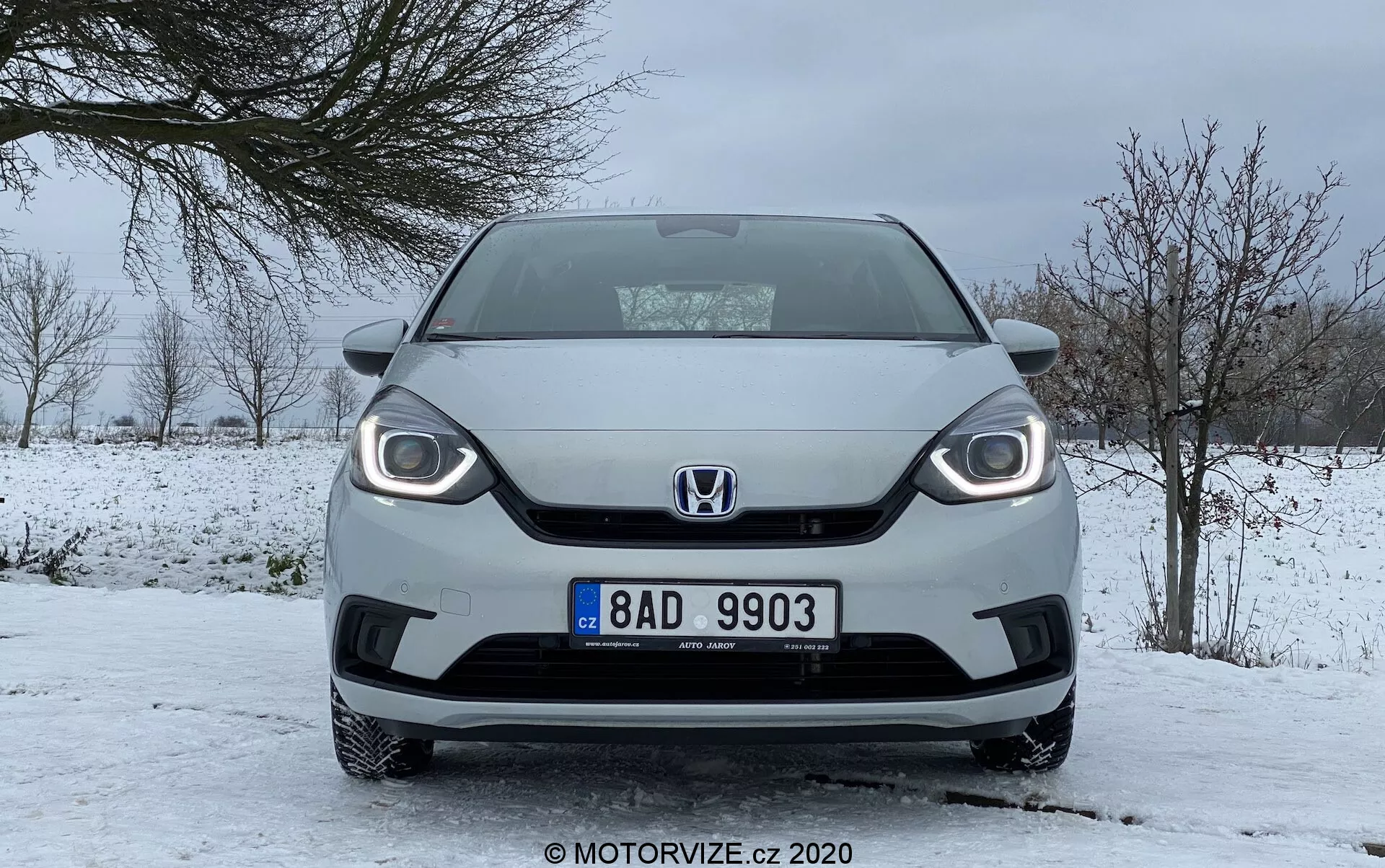 Vista frontal de um Honda Jazz 2020 em condições de inverno, apresentando design distinto da grade, faróis visíveis com luzes diurnas de LED e placa de licença central, estacionado em uma estrada coberta de neve com céu nublado e árvores sem folhas.