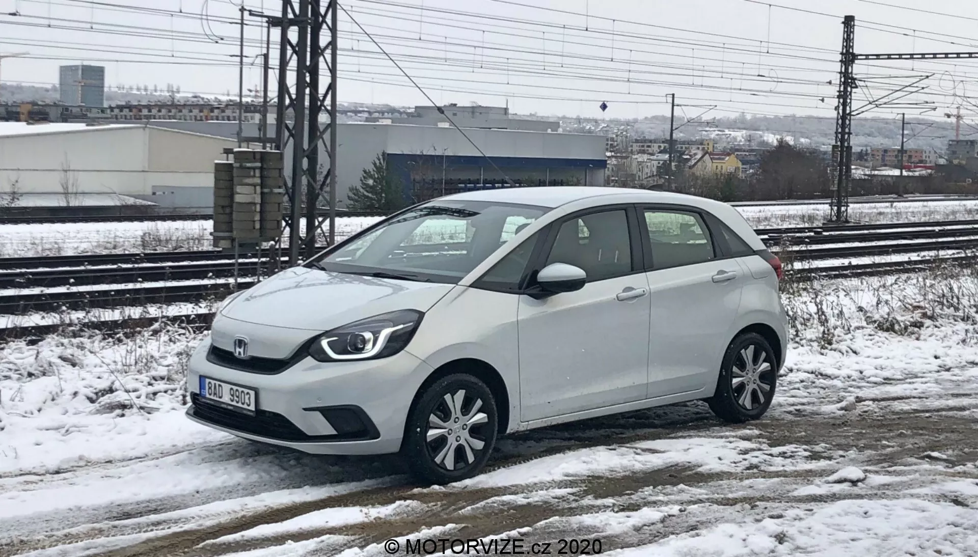 Um Honda Jazz 2020 branco, um carro hatchback subcompacto, capturado de uma perspectiva de altura baixa a média, mostrando tanto a frente quanto o lado direito. O carro está estacionado em uma superfície nevada e molhada, com neve visível no veículo e no chão, indicando condições climáticas frias. Trilhos de trem e estruturas industriais são visíveis ao fundo sob um céu nublado. O carro apresenta uma grade frontal principal com o emblema da marca, faróis de halogênio, rodas de liga leve com vários raios, espelhos laterais com indicadores de direção integrados e uma antena curta no teto traseiro.