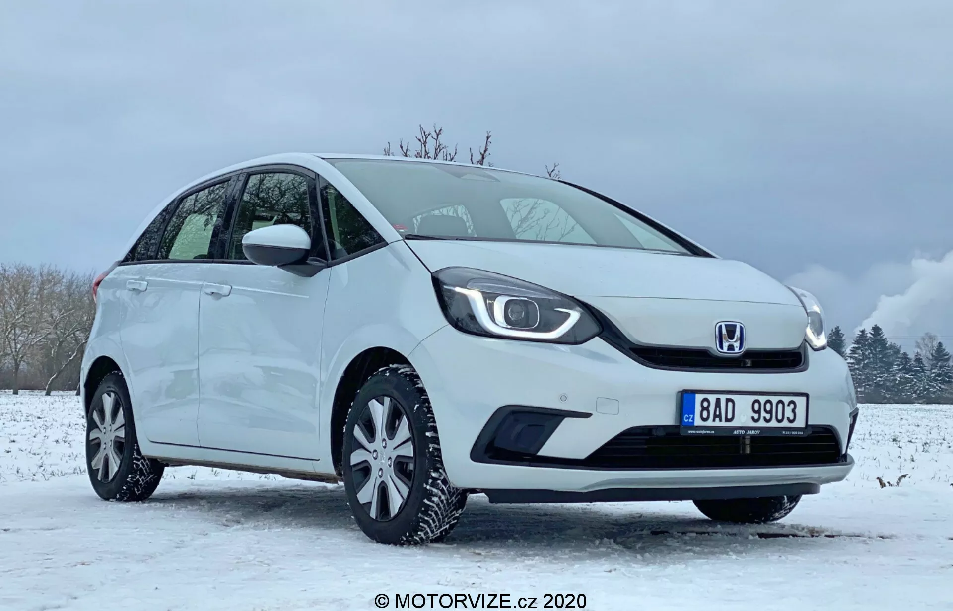 A imagem mostra um Honda Jazz 2020 compacto hatchback branco posicionado em um cenário de inverno com chão coberto de neve e céu parcialmente nublado. O carro é visto de um ângulo frontal lateral focado no lado do motorista, destacando a grade distintiva com o logotipo no centro, faróis elegantes de LED e para-choque dianteiro liso com carcaça de luzes de neblina integrada. O perfil lateral exibe uma linha de teto inclinada, rodas de liga leve com raios múltiplos e espelhos laterais e maçanetas da cor do corpo. A condição impecável do veículo sugere que pode ser uma imagem promocional ou de revisão capturada em 2020 pela MOTORVIZE.cz.