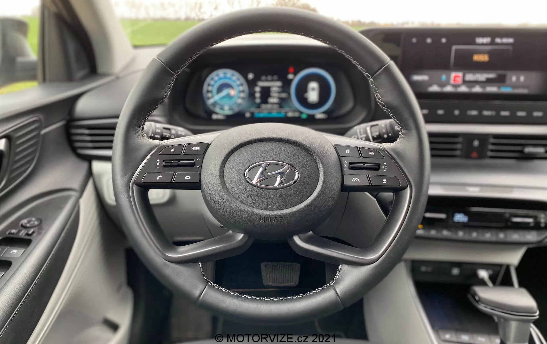 Vista interior do Hyundai i20 2020 da perspetiva do condutor, mostrando um volante moderno de três raios com o logótipo Hyundai, botões multifuncionais e um painel de instrumentos totalmente digital exibindo velocímetro, tacómetro e ecrã de informações do condutor. O painel de instrumentos apresenta um display central de infotainment com opções de menu, indicando uma integração tecnológica avançada e uma interface amigável para o utilizador. O design interior é moderno, com linhas limpas e materiais de qualidade visíveis na consola central e na porta do lado do passageiro.
