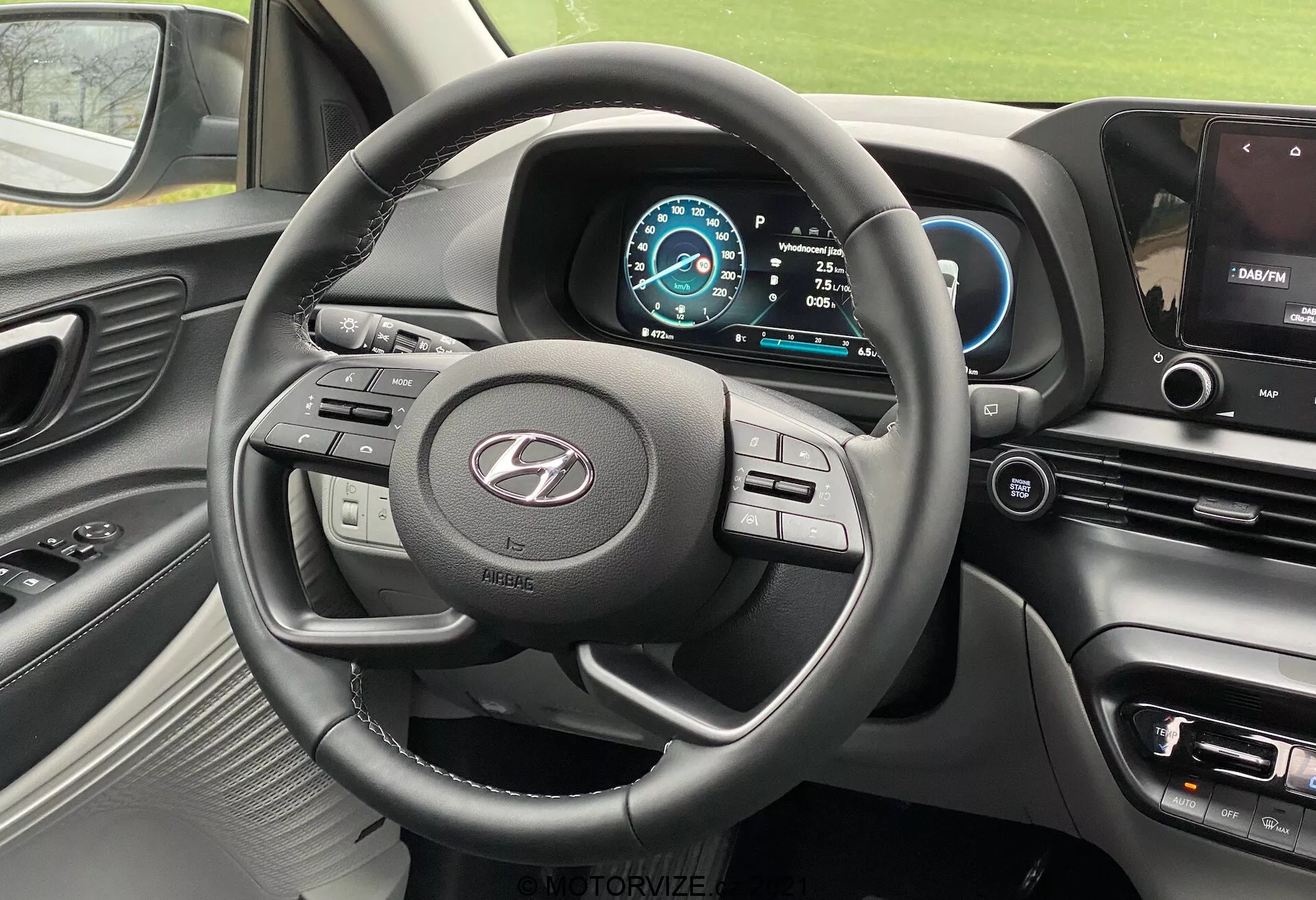 Vista interior do Hyundai i20 2020 a partir do assento do motorista, mostrando o volante de três raios com múltiplos botões de controle, o logotipo do fabricante em destaque, painel de instrumentos digital com esquema de cores azul e branco, velocímetro ajustado para 0 km/h, seleção de marcha em Park, parte do console central com tela de infoentretenimento exibindo um mapa de navegação, botões de controle de clima e configurações do veículo, painel de toque suave com detalhes metálicos ao redor das saídas de ar e do volante, e vista parcial da saída de ar do lado do passageiro.