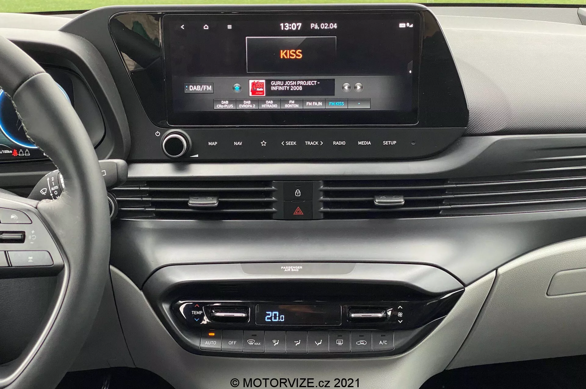 Fotografia do painel interior de um carro moderno a partir da perspetiva do banco do condutor, destacando a consola central com um grande ecrã de infoentretenimento, saídas de ar integradas, botão de luzes de emergência e um elegante painel de controlo de climatização com ecrã digital de temperatura e botões sensíveis ao toque. O design do painel apresenta um estilo sofisticado com materiais de alta qualidade e acentos metálicos. A borda do volante com botões de controlo integrados é parcialmente visível à esquerda.