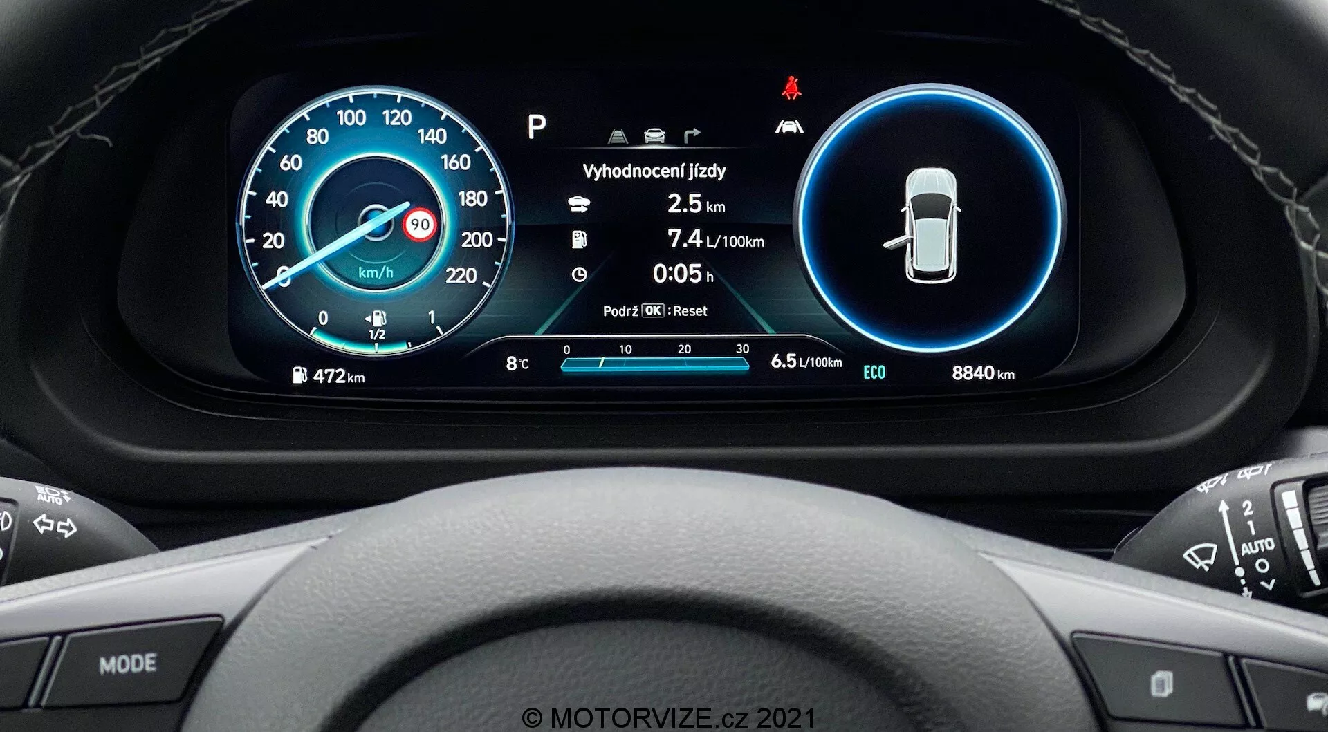 Perspectiva do banco do motorista do Hyundai i20 2020, mostrando uma visão detalhada do painel de instrumentos digital situado atrás do volante. O design moderno inclui dois principais displays circulares: um velocímetro digital e analógico à esquerda e indicadores de características de segurança à direita. No centro, são exibidas informações adicionais como temperatura, odômetro, economia de combustível e seleção de marchas. Na periferia, vemos a quilometragem total (8840 km), indicador de combustível e vários ícones do sistema. O volante multifuncional com botões de mídia e controle de cruzeiro é parcialmente visível. Na parte inferior, há uma marca d'água 'MOTORVIZE.cz 2021'.