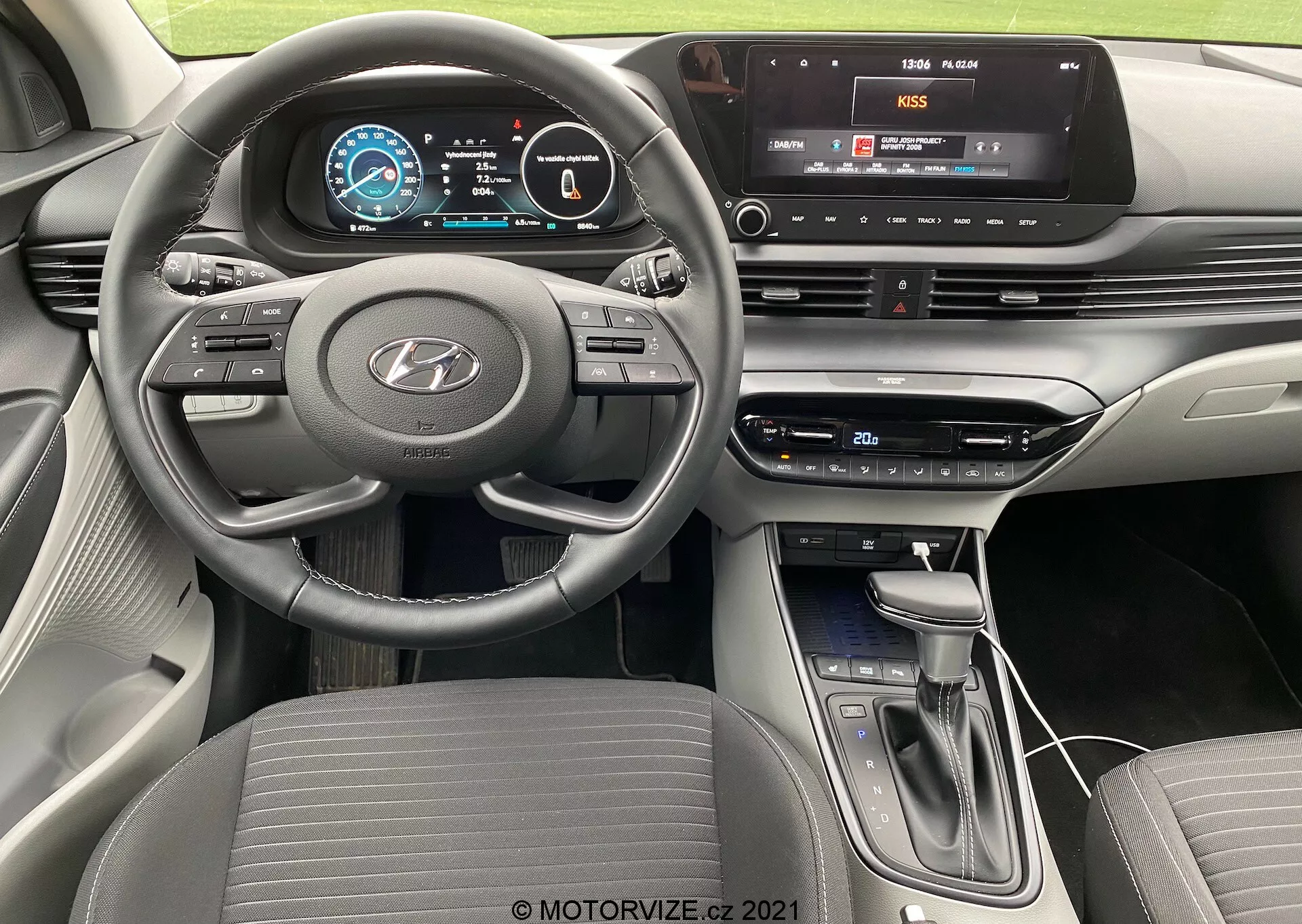Vista do interior do Hyundai i20 2020 a partir da perspectiva do motorista, apresentando volante de três raios com fundo plano e controles montados para áudio, controle de cruzeiro e comunicação mãos-livres, um painel de instrumentos totalmente digital com gráficos coloridos e um console central com sistema de infoentretenimento com tela sensível ao toque e conectividade multimídia, botões e botões físicos para controle do clima, alavanca de câmbio moderna e design contemporâneo em camadas do painel com esquema de cores bicolores.