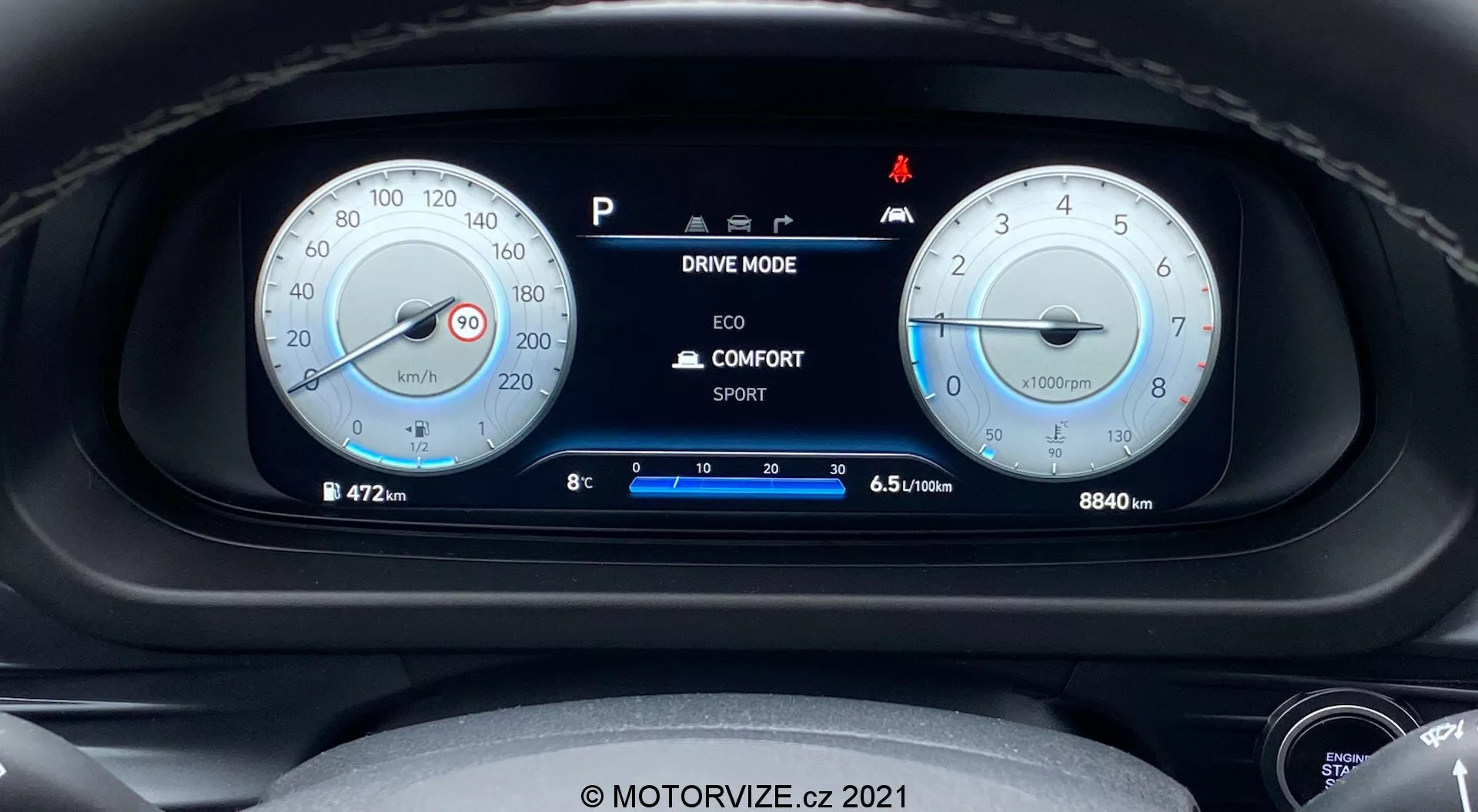 Vista aproximada do painel de instrumentos do Hyundai i20 2020 a partir da perspectiva do motorista, com dois medidores analógicos redondos principais e um display digital central. O medidor à esquerda mostra um velocímetro marcado até 260 km/h, enquanto o da direita é um conta-giros com uma linha vermelha a 6500 rpm. A tela digital central mostra opções de modo de condução, temperatura externa, posição da marcha e eficiência de combustível. O odômetro marca 8840 km. Várias luzes indicadoras são visíveis, incluindo uma para a função de partida/parada automática. A imagem é nítida e bem iluminada, com ligeiro suavizado ao redor das bordas dos medidores. Inclui marca d'água 'MOTORVIZE.cz 2021'.