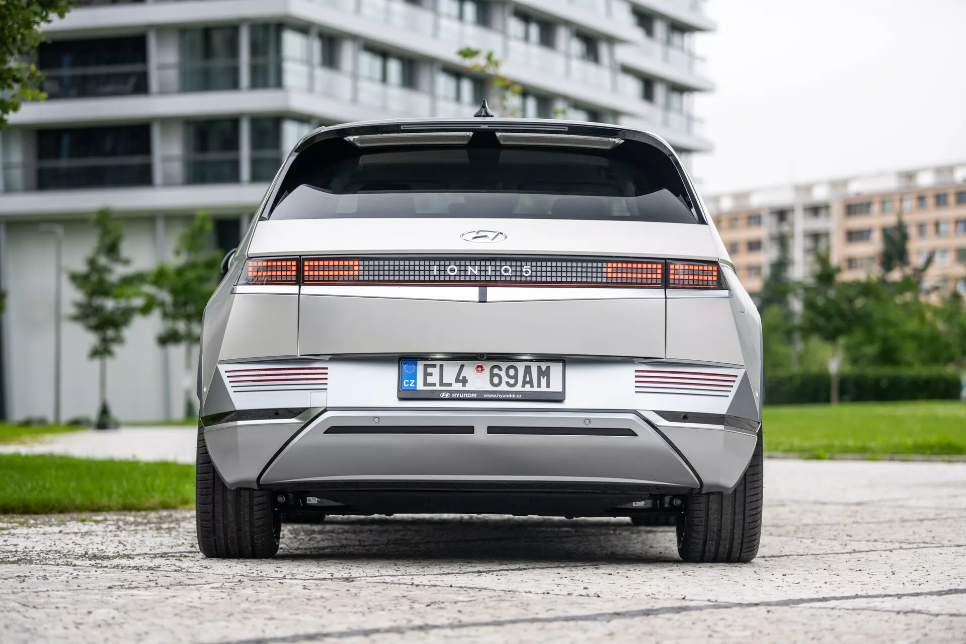 Vista traseira do Hyundai Ioniq 5 2WD 2021, destacando uma barra luminosa traseira distinta de largura total, logotipo do fabricante centralizado, parte traseira plana com linhas horizontais nítidas, para-choque traseiro minimalista com tiras reflexivas e placa de formato europeu. O veículo está centrado em uma superfície de asfalto com folhagem e edifícios modernos ao fundo, sob uma iluminação nublada.