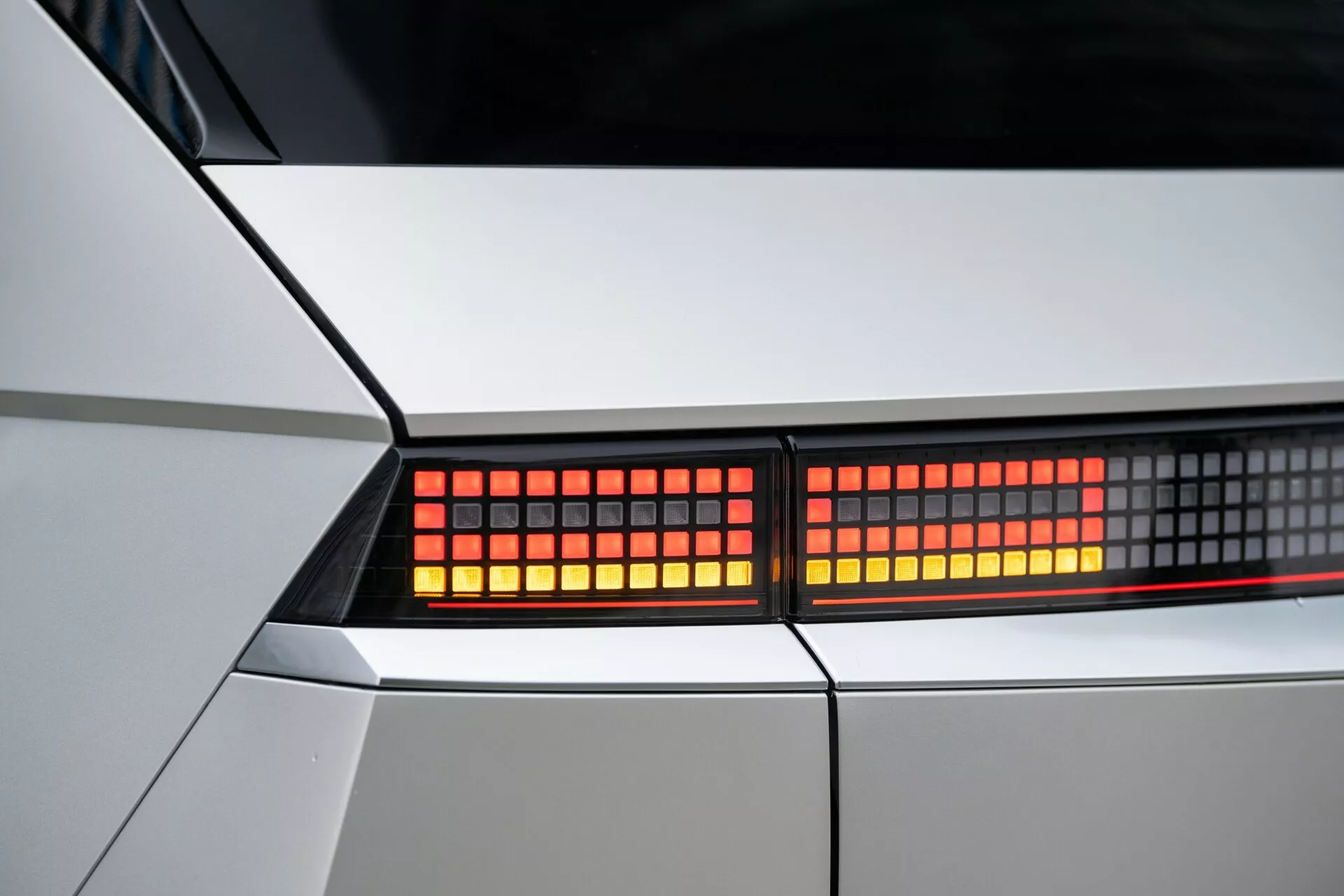 Imagem em close da montagem da luz traseira do Hyundai Ioniq-5 2021 2WD. Foco nos LEDs vermelhos e pixelizados em um enquadramento estreito, destacando a carroceria geométrica do carro em branco ou prata claro. Vista detalhada dos elementos de design moderno e da tecnologia de iluminação.