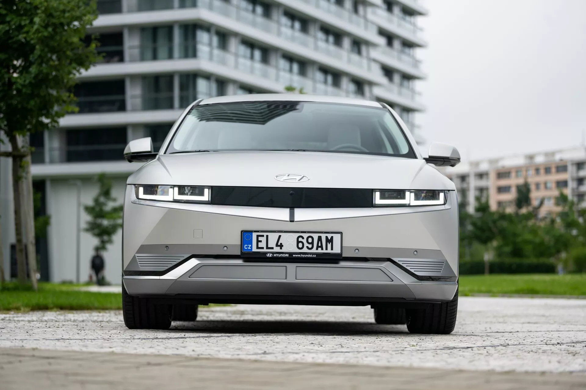 A imagem retrata um Hyundai Ioniq 5 2WD 2021, destacando sua parte frontal de um ângulo baixo. A fotografia capta a grade moderna, os faróis LED avançados e o para-choque aerodinâmico com recortes detalhados. O carro, um modelo elétrico, possui uma grade fechada típica de veículos elétricos, uma faixa de luz sob o capô e sensores para sistemas avançados de assistência ao motorista. O capô, adornado com o emblema do fabricante, é limpo e suave. O pano de fundo urbano destaca a adequação do carro para a vida na cidade.