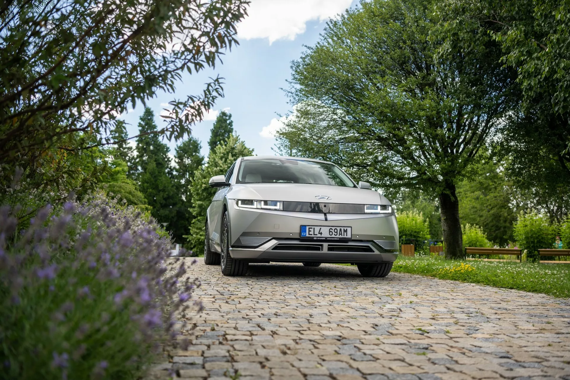 Foto tirada de baixo ângulo de um Hyundai Ioniq 5 2WD 2021 estacionado ao ar livre sobre uma superfície de paralelepípedos. A câmera foca na frente do carro, incluindo a grade, os faróis esquerdos e parte do capô e para-brisa, com um fundo de árvores verdes, arbustos e flores roxas sob abundante luz natural.