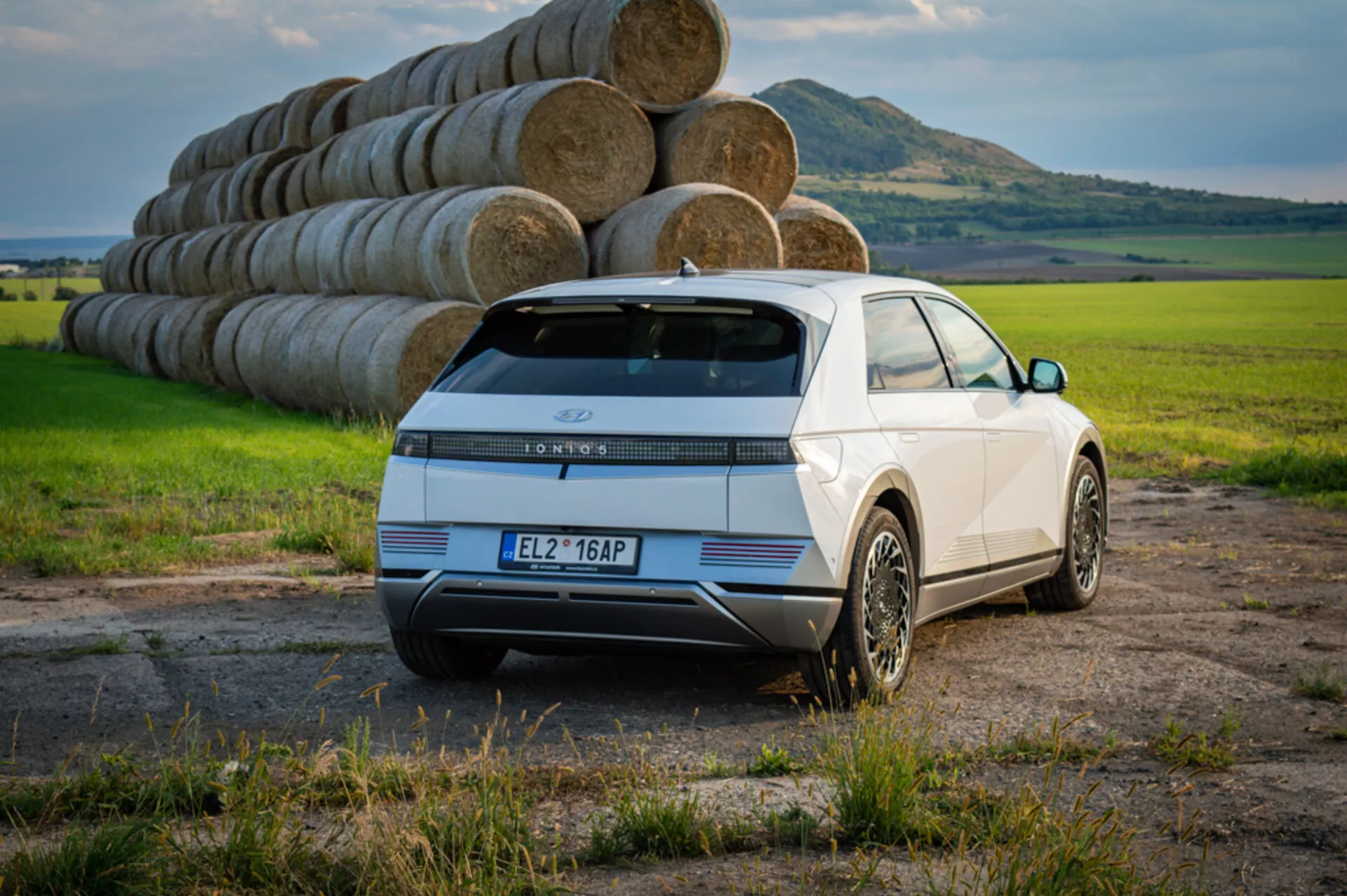 SUV elétrico Hyundai Ioniq 5 de 2021 em branco, exibindo visão traseira e lateral com lanternas traseiras LED, rodas de liga leve estilosas e sensores contra um fundo de campo aberto e fardos de feno sob céu claro diurno