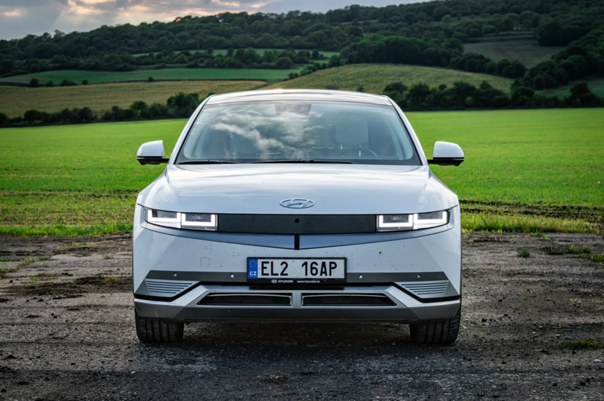 Vista frontal do veículo elétrico Hyundai Ioniq 5 de 2021 em cascalho contra campo verdejante, com faróis LED finos e design aerodinâmico sem grade tradicional.