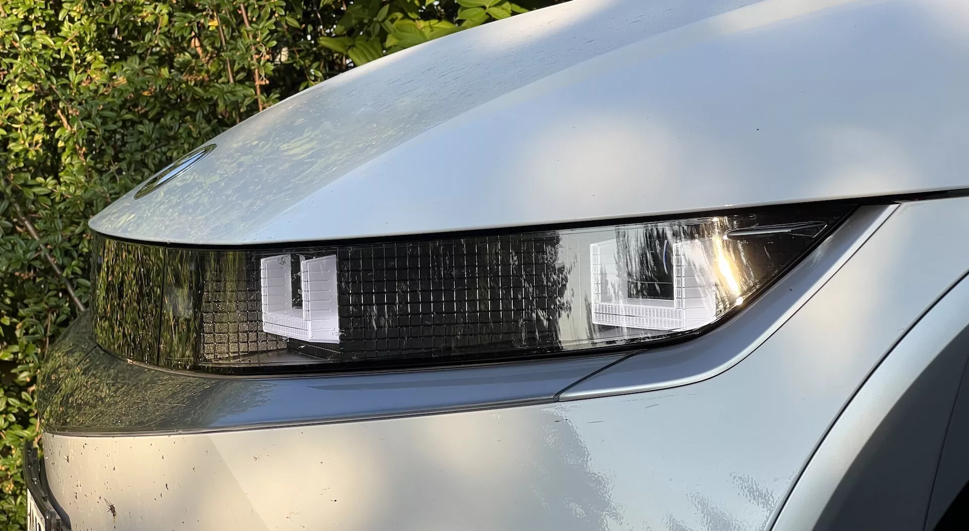 Vista de perto do farol dianteiro esquerdo do Hyundai Ioniq-5 2021, exibindo iluminação LED ou HID com segmentos em formas geométricas, uma cobertura clara e brilhante e reflexos na pintura metálica com sujeira visível, indicando uso.