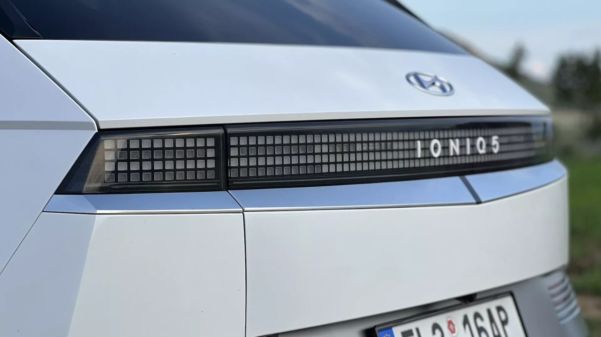 Vista detalhada da traseira de um Hyundai Ioniq 5 de 2021, destacando o emblema da marca, o nome do modelo e a barra de luz LED avançada, contra um fundo exterior desfocado.