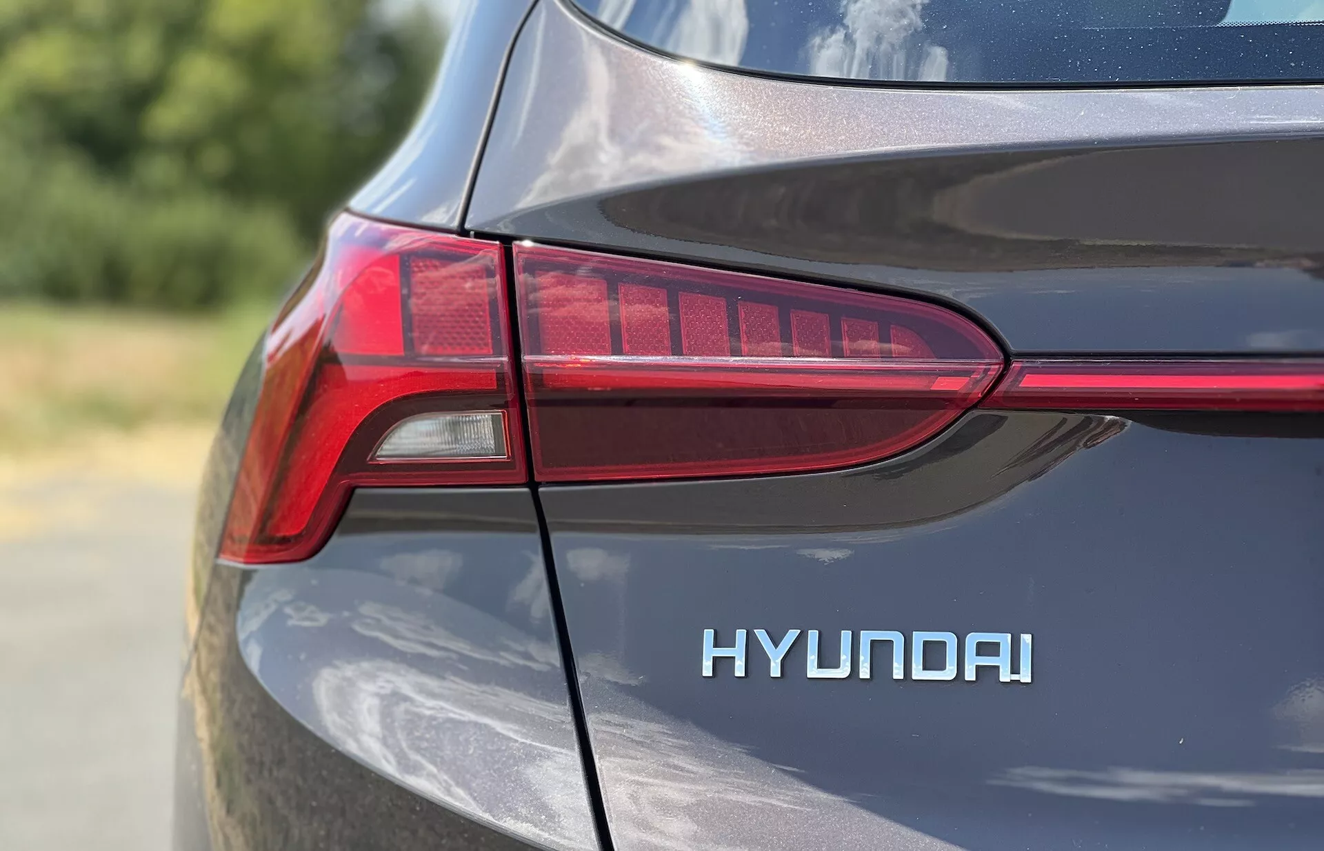 Close-up da quarta traseira do lado do motorista de um Hyundai Santa-Fe 2020, focando no design em LED em forma de T da lanterna traseira e no emblema 'HYUNDAI' destacado. O acabamento metálico da pintura reflete a luz natural do dia, destacando as curvas e linhas do design do carro em uma foto apertada e detalhada com pouca profundidade de campo.