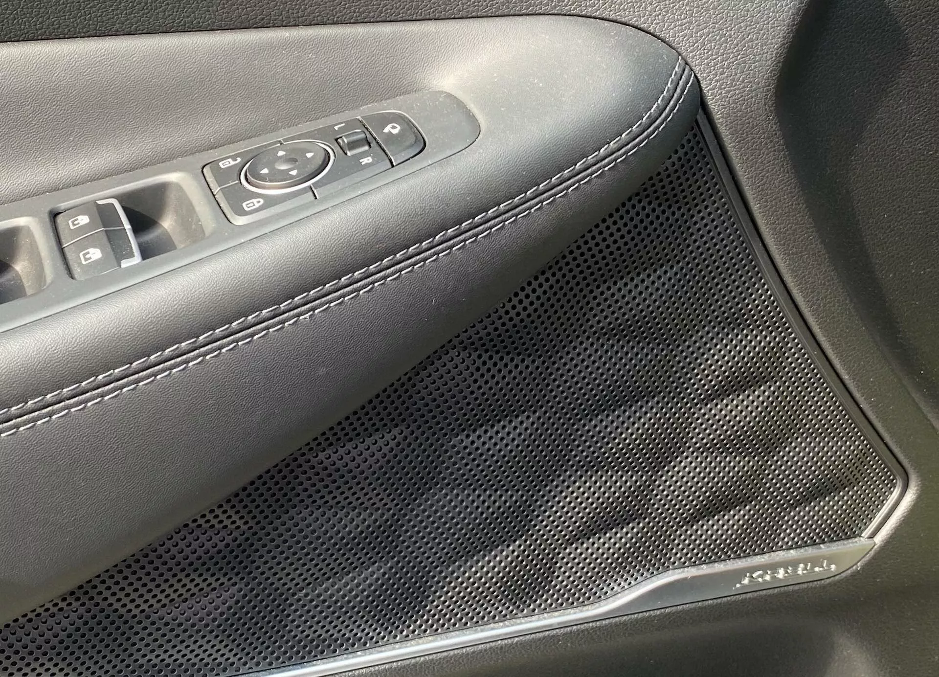 Close-up do painel interno da porta de um Hyundai Santa Fe 2020, destacando a área com botões de controle dos vidros e espelhos elétricos, assim como uma grade de alto-falante com padrão de colmeia. O apoio de braço é estofado em material escuro com costura branca. Um detalhe cromado adorna a grade do alto-falante, indicando características avançadas de áudio e design estético.