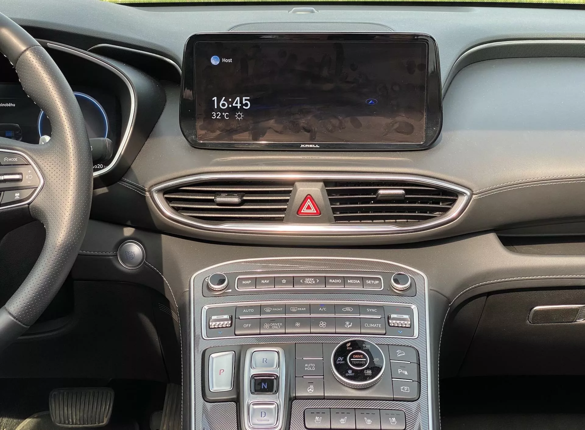 Close-up, vista frontal do console central e parte do painel de instrumentos de um carro moderno, incluindo a tela de infoentretenimento mostrando 16:45 e 32°C, saídas de ar aerodinâmicas, um botão de luzes de emergência, uma combinação de controles sensíveis ao toque e botões físicos com detalhes metálicos, um botão rotativo para controle do sistema de infoentretenimento e navegação e um seletor de modo de condução, tudo sugerindo um interior elegante e moderno com funções avançadas, como controle de clima, aquecimento/resfriamento de assentos e vários modos de condução. O painel de instrumentos desfocado e a visão parcial do volante à esquerda implicam um veículo de alta qualidade projetado para mercados com direção à esquerda.