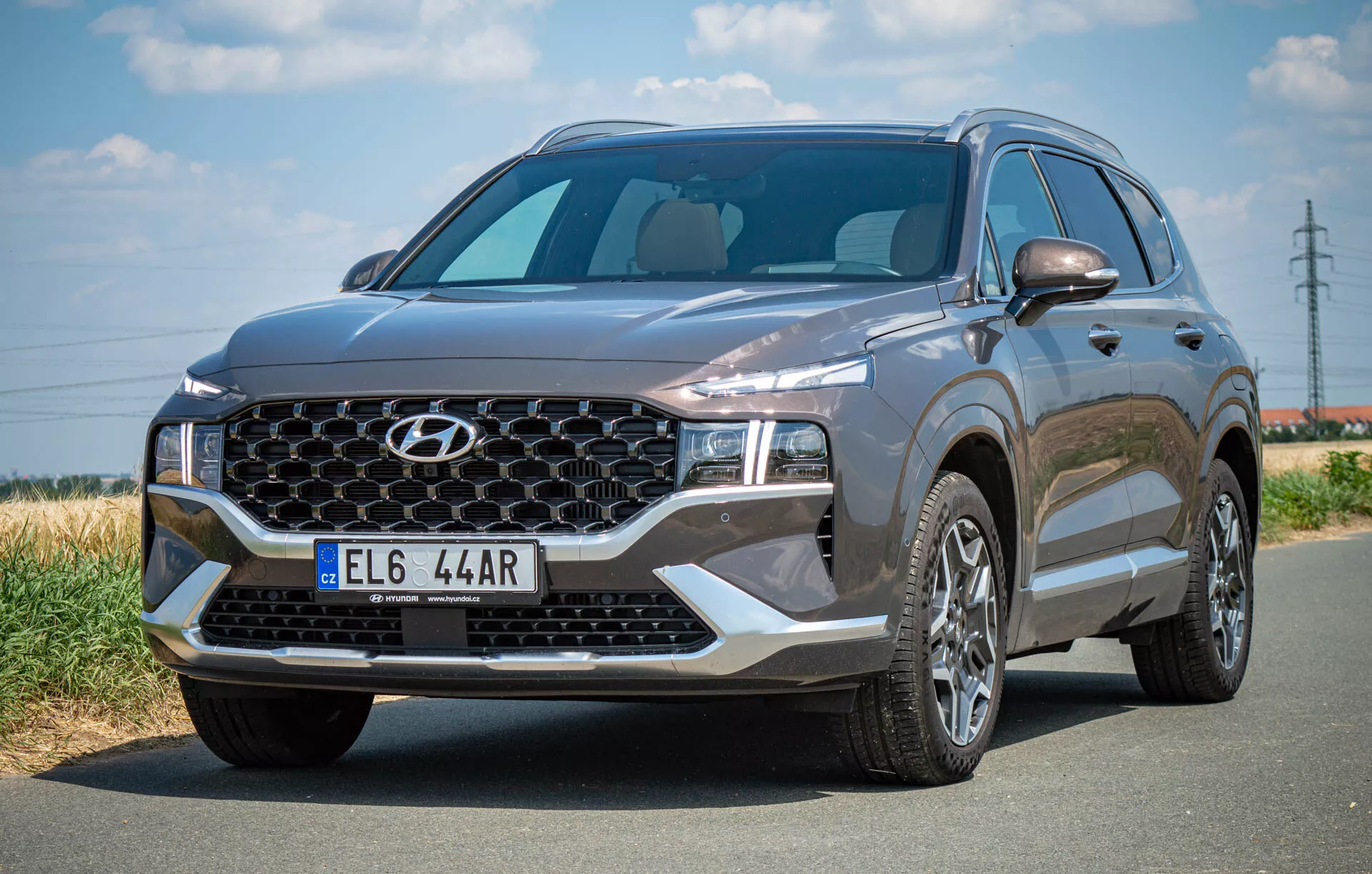 A imagem mostra um SUV Hyundai Santa Fe 2020 estacionado em uma estrada de asfalto, capturado de um ângulo frontal esquerdo de três quartos. Destaca-se a grande grade com um padrão específico da marca e logotipo central, os elegantes faróis de LED e o capô esculpido. O para-brisa é amplo e oferece boa visibilidade, e o perfil lateral revela rodas de liga leve multirraiadas e o espelho lateral do motorista com piscas integrados potencialmente. O veículo de cor escura transmite uma aparência sofisticada com seus elementos de design modernos, incluindo possíveis sensores ou câmeras frontais.