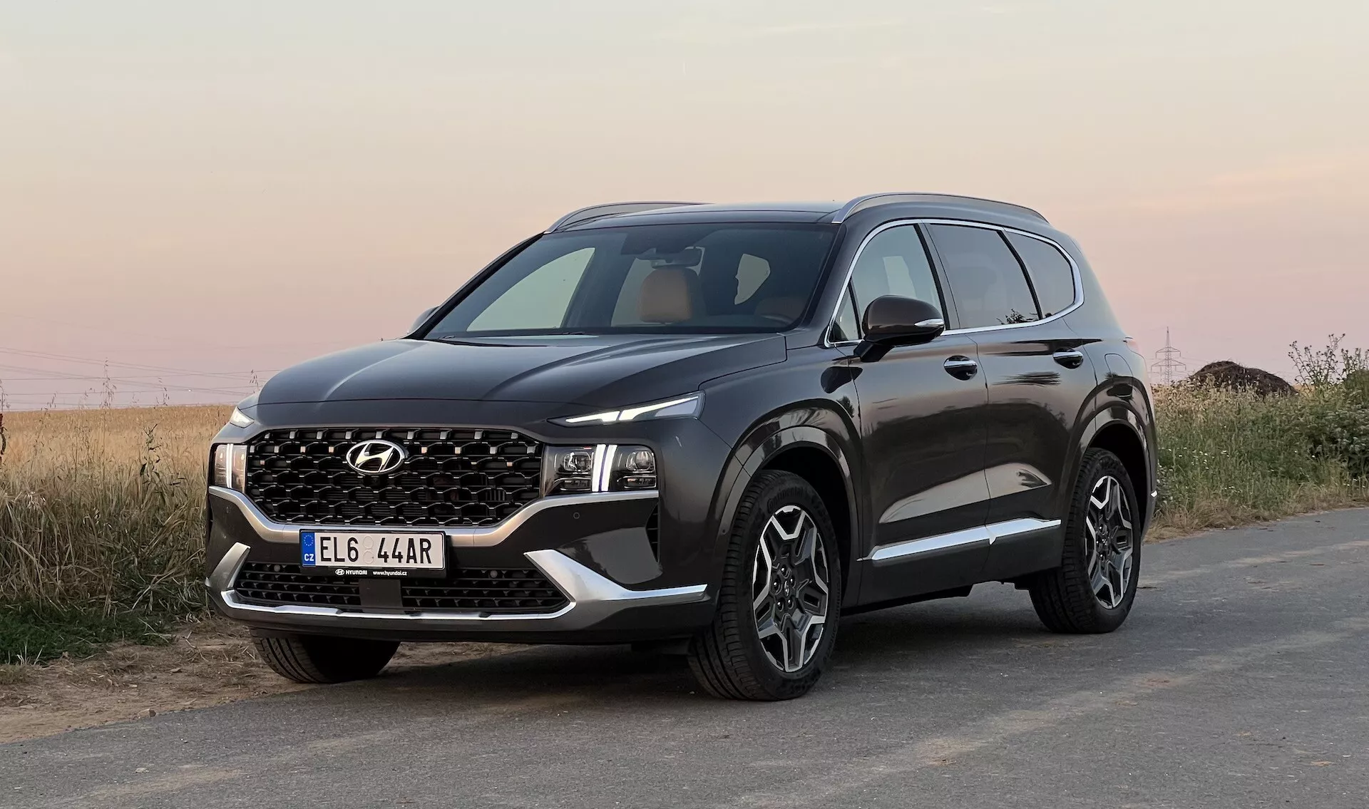 Imagem de um SUV Hyundai Santa Fe 2020 estacionado em uma superfície de asfalto, tirada de um ângulo baixo com vista frontal de três quartos, exibindo a grade dianteira, faróis estreitos de LED, para-choque com luzes de neblina e placa de proteção falsa, capô com vincos angulares, rodas grandes de liga leve multirraios, revestimento protetor preto nos arcos das rodas e na parte inferior das portas, espelho lateral com sinais de seta integrados e janelas com moldura cromada. O fundo apresenta uma paisagem natural indefinida sob um céu aberto.