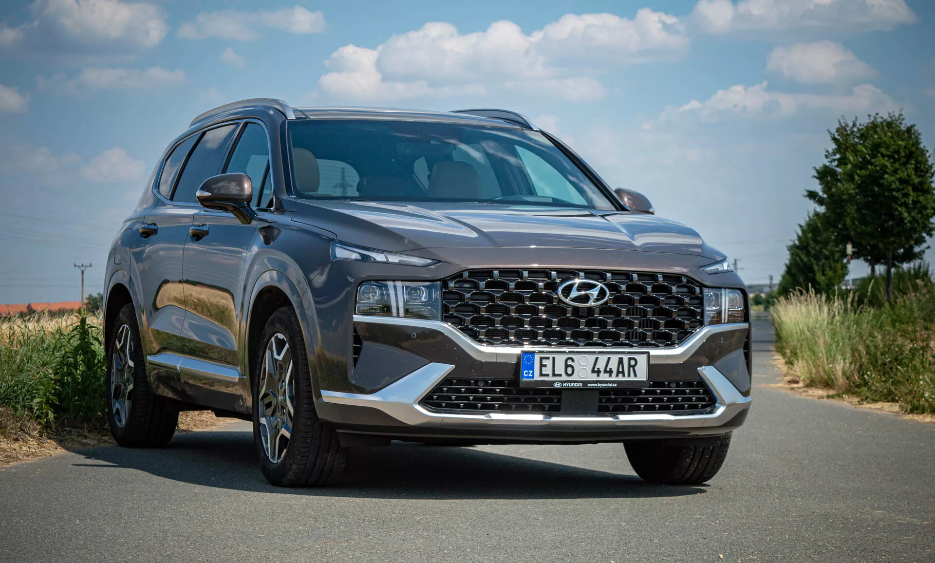 Imagem de um SUV Hyundai Santa-Fe 2020 estacionado em uma estrada pavimentada sob um céu claro com vegetação ao fundo. A foto foi tirada de uma perspectiva de três quartos frontal, mostrando a fachada frontal do veículo e o perfil lateral esquerdo. O SUV possui uma grande grade com padrão entrelaçado com o emblema da Hyundai, faróis angulares elegantes em LED ou HID, rodas de liga leve de vários raios, uma carroceria moderna e aerodinâmica com linhas suaves, retrovisores laterais com sinais de direção integrados, vidros escurecidos, cor cinza escuro, placa dianteira e parece estar em perfeitas condições, sugerindo que é novo ou bem mantido.